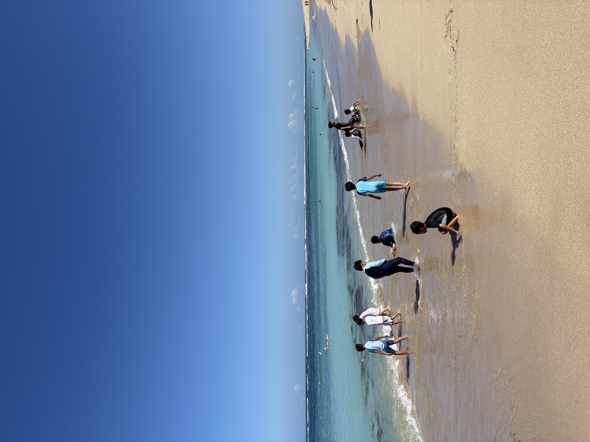 The image shows a beach in Nusa Dua, Indonesia. The sky is clear and blue. The sand is light brown, and the water is turquoise and clear. There are several people on the beach, mostly children. Some are walking along the shoreline, while others are playing in the sand and water. The waves are small and gentle.