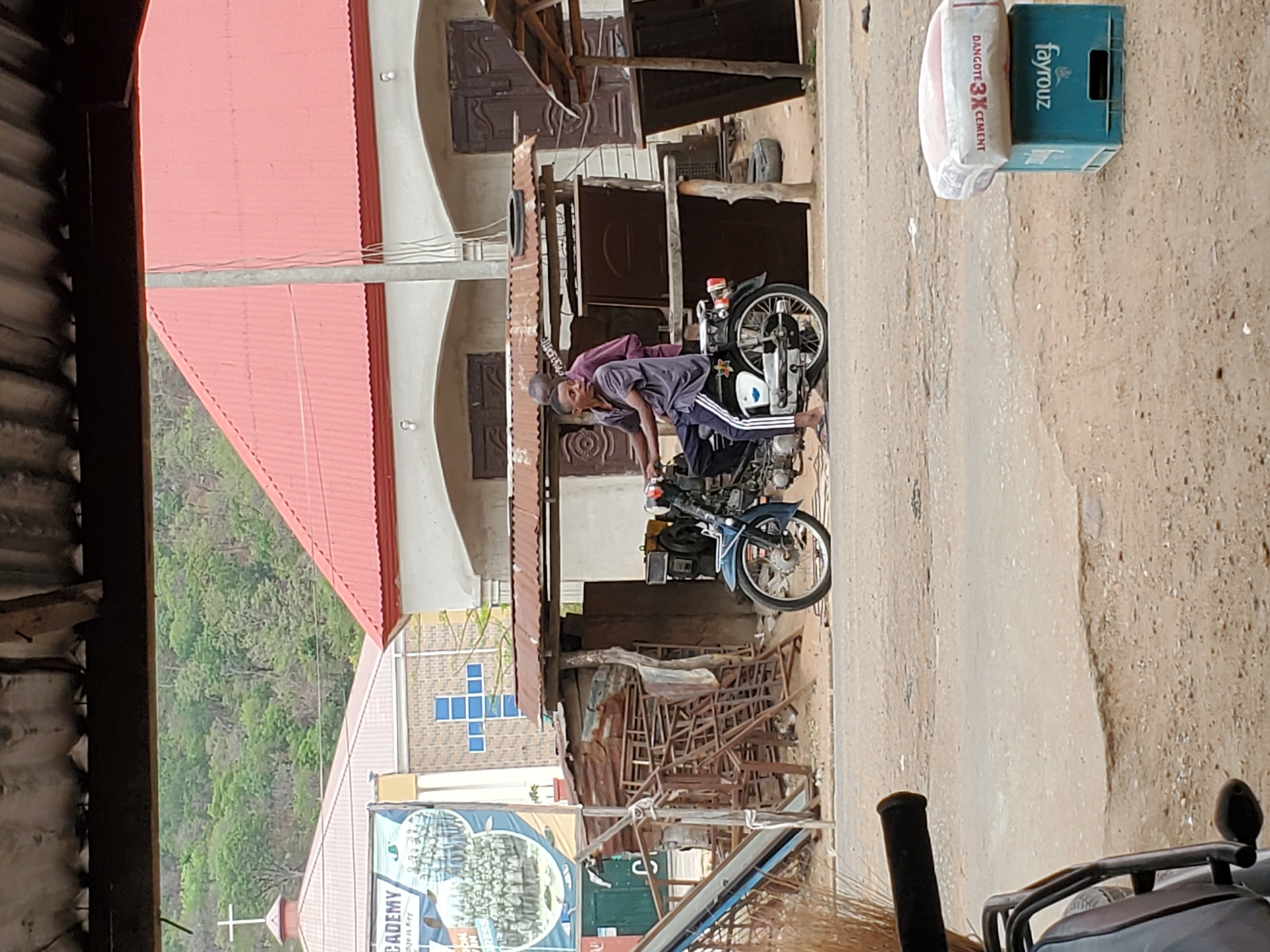A man straddles a motorcycle in a street scene in Mararaba, Nigeria. He is wearing a dark shirt and pants with white stripes. On the side of the road is a bag of Dangote 3X cement and a Fayrouz crate. A building with a red roof is visible to the left, as well as another building with a blue cross design. Discarded metal and other debris is piled up near the buildings. The street is dirt-covered. A tree covered hillside can be seen.
