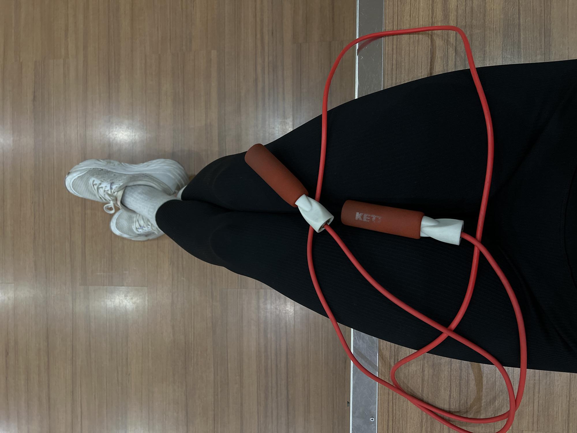 The image shows a close-up, overhead view of a person's legs and a jump rope. The person is wearing black leggings and white athletic socks with white sneakers. The jump rope, with red cord and orange handles, is coiled on top of the leggings. The background is a light brown wooden floor. There is a metallic strip running horizontally across the lower portion of the image, suggesting a gym or fitness studio setting. The image provides no information or context to determine a specific location within Denpasar, Indonesia.