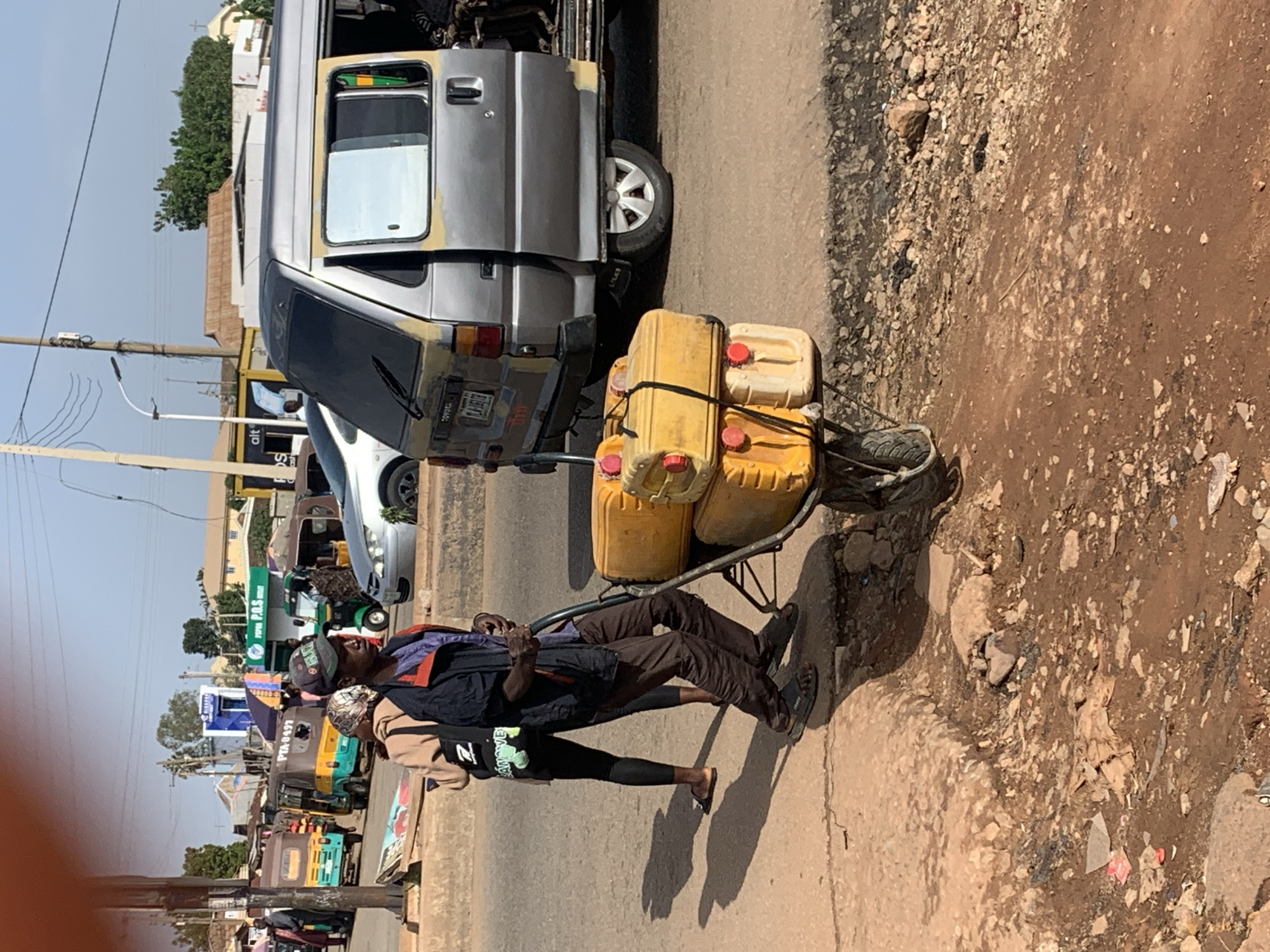The image depicts a street scene in Gura Top, Nigeria, where a man is pushing a wheelbarrow loaded with several yellow jerrycans. The man, wearing brown pants and a dark jacket, is positioned on the right side of the frame, actively engaged in moving the wheelbarrow. He is wearing sandals and a cap. Behind him, a person in a brown jacket and black leggings is walking away from the viewer. In the background, a silver van with its side door open is parked on the road. Other vehicles, including what appear to be auto-rickshaws or similar small transport vehicles, are visible further down the street. The environment appears to be a busy urban area with a mix of buildings, stalls, and a dirt road with some debris. The lighting suggests it is daytime, with shadows cast on the ground.