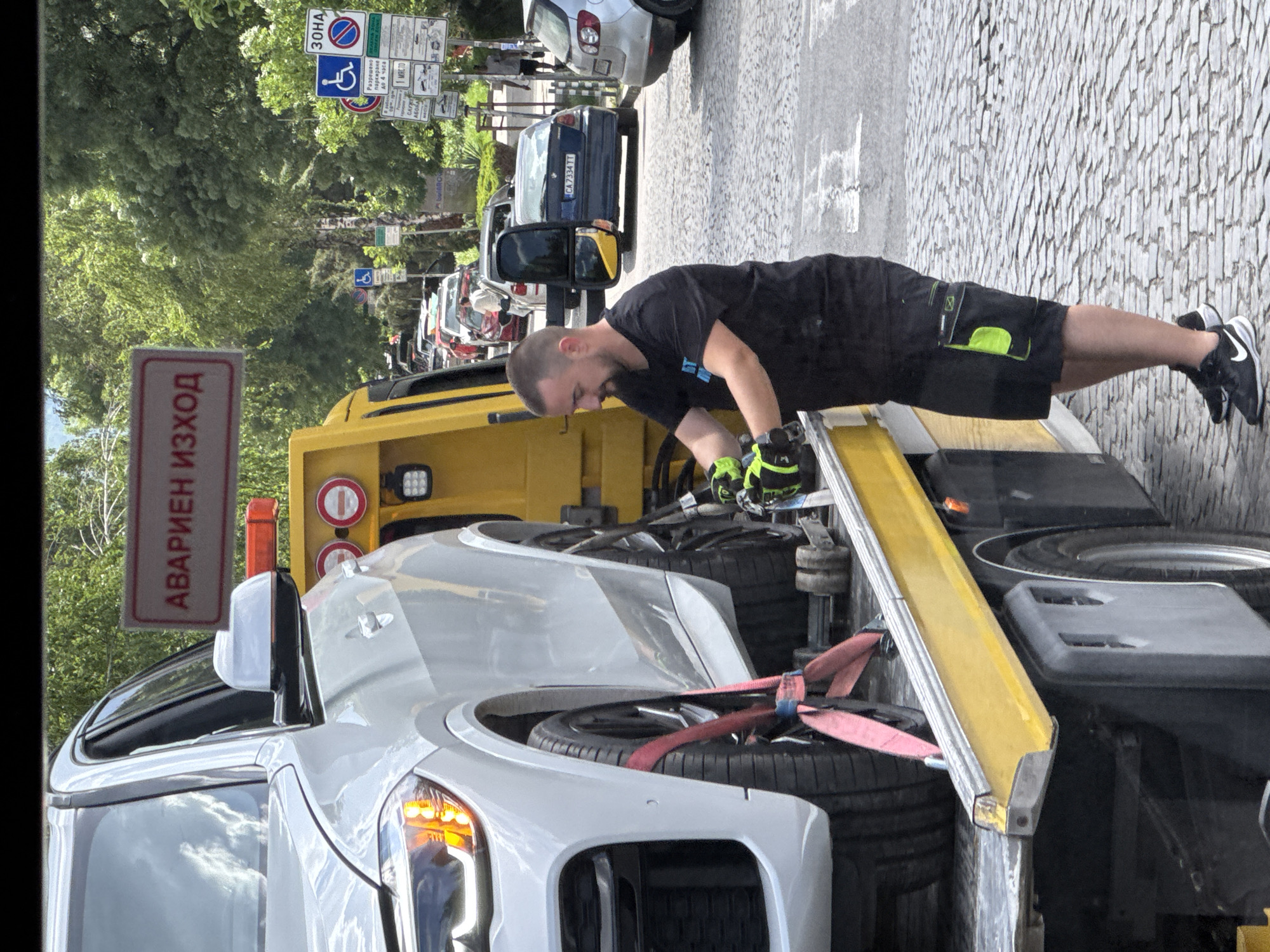 The image shows a man in a black t-shirt, black shorts with green accents, and black sneakers working on a white car that is secured on a yellow tow truck. He is wearing gloves with green accents and appears to be inspecting or adjusting something on the car. The tow truck has an "АВАРИЕН ИЗХОД" (Emergency Exit) sign. The background features a street with other parked cars, trees, and several road signs, including one indicating a disability parking zone and others with text in Bulgarian. The pavement is made of cobblestones. Based on the style of signs and architecture, the location is likely Sofia, Bulgaria.