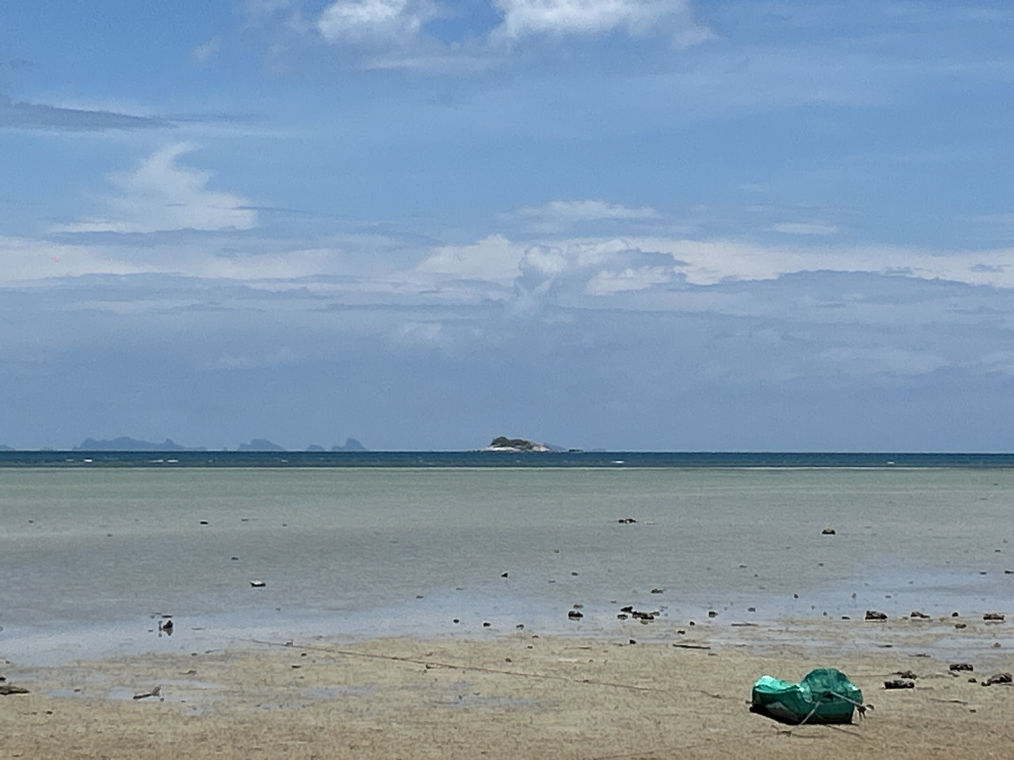 The image shows a coastal scene in ตำบลเกาะพะงัน, Thailand. The tide is low, revealing a wide expanse of shallow water and sandy beach. There are rocks and other small debris scattered across the exposed seabed. In the distance, there are land masses, including a small island. The sky is blue with patchy clouds. In the foreground, there is a green, possibly discarded, object on the sand.