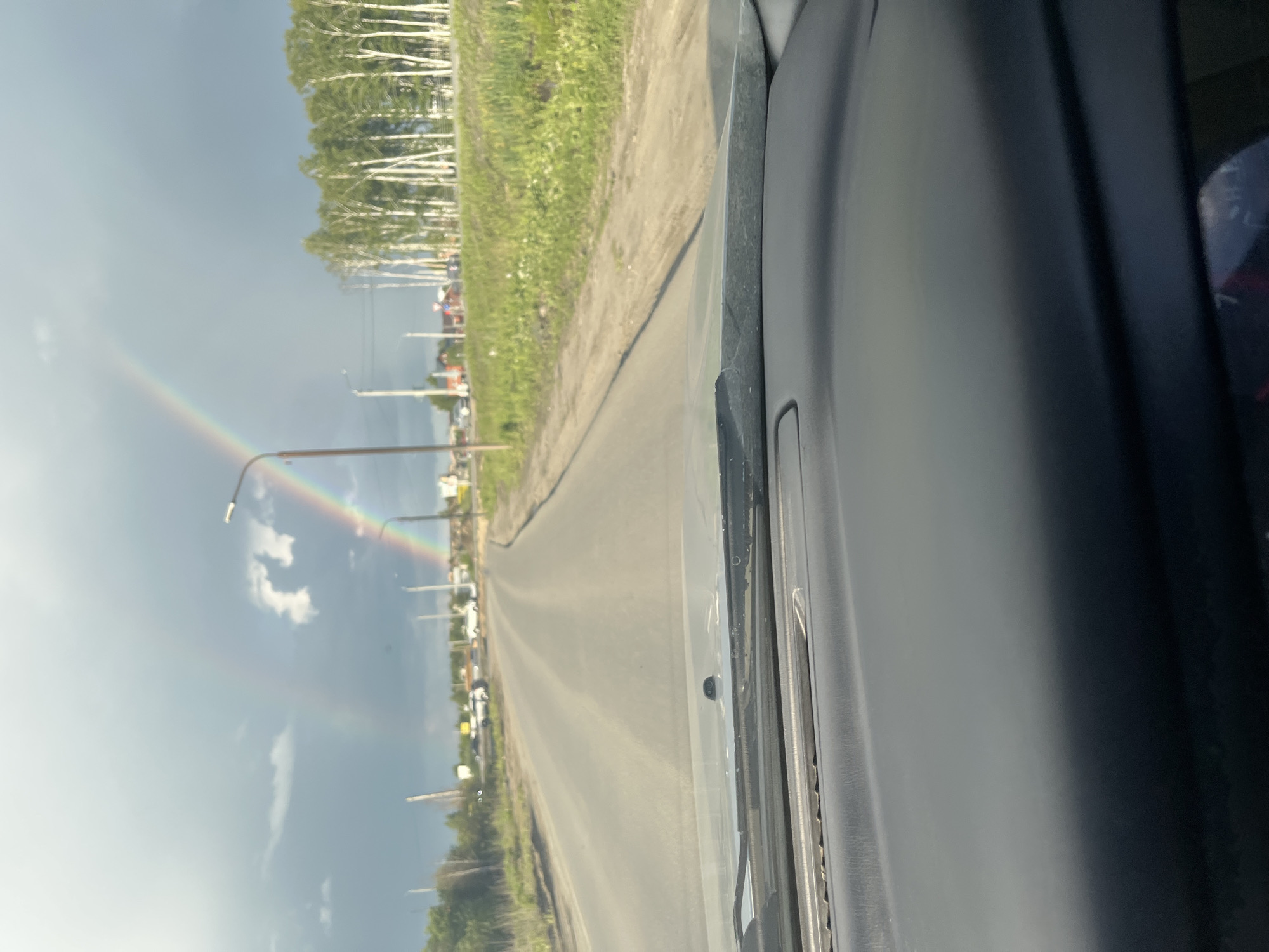 The image is taken from the driver's perspective inside a vehicle, looking out at a road. A double rainbow is visible in the overcast sky. The road appears to be a two-lane asphalt surface leading into the distance, with a grassy verge and a line of birch trees on the right side of the road. In the distance, there are some buildings and streetlights. The foreground shows the dashboard of the vehicle. The scene is consistent with a suburban or rural area. The location is identified as Prigorodny, Russia.