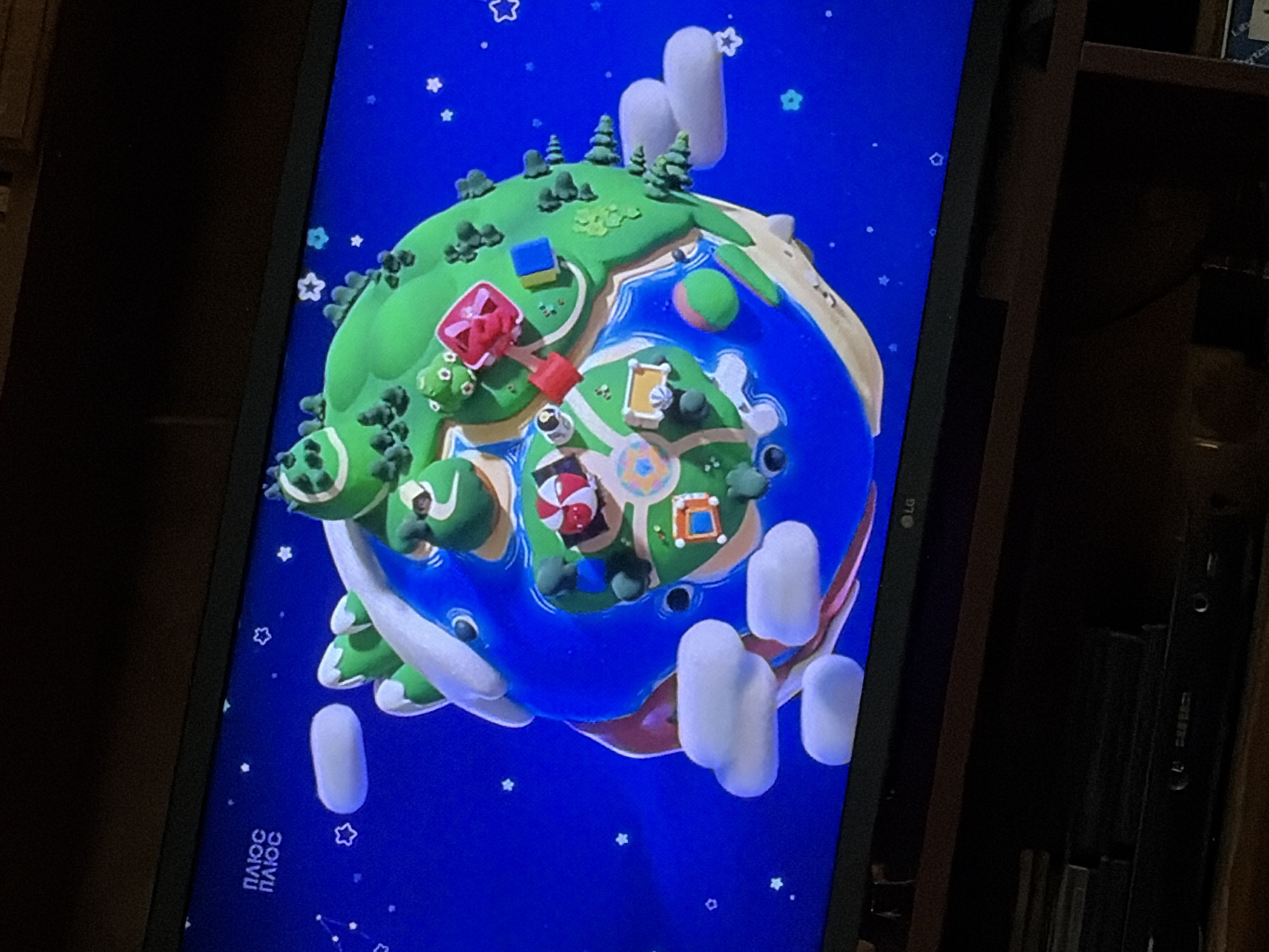 The image shows a television screen displaying a 3D animated scene of a cartoon planet. The planet is round and has a blue ocean surface with green landmasses dotted with trees and small buildings. There are white clouds floating around the planet, and a starry blue background with small white star shapes. The planet has various cartoonish elements, including what appear to be miniature houses, roads, and recreational areas. In the upper left corner of the screen, there is text in a foreign language, likely Cyrillic, which reads "плюс плюс" (plus plus). The television is a flat-screen model, and the bottom of the screen shows an LG logo. Below the television, there is a dark shelf with stacks of media cases and an electronic device. The overall scene is a digital illustration presented on a television. The location is not specified in the image.