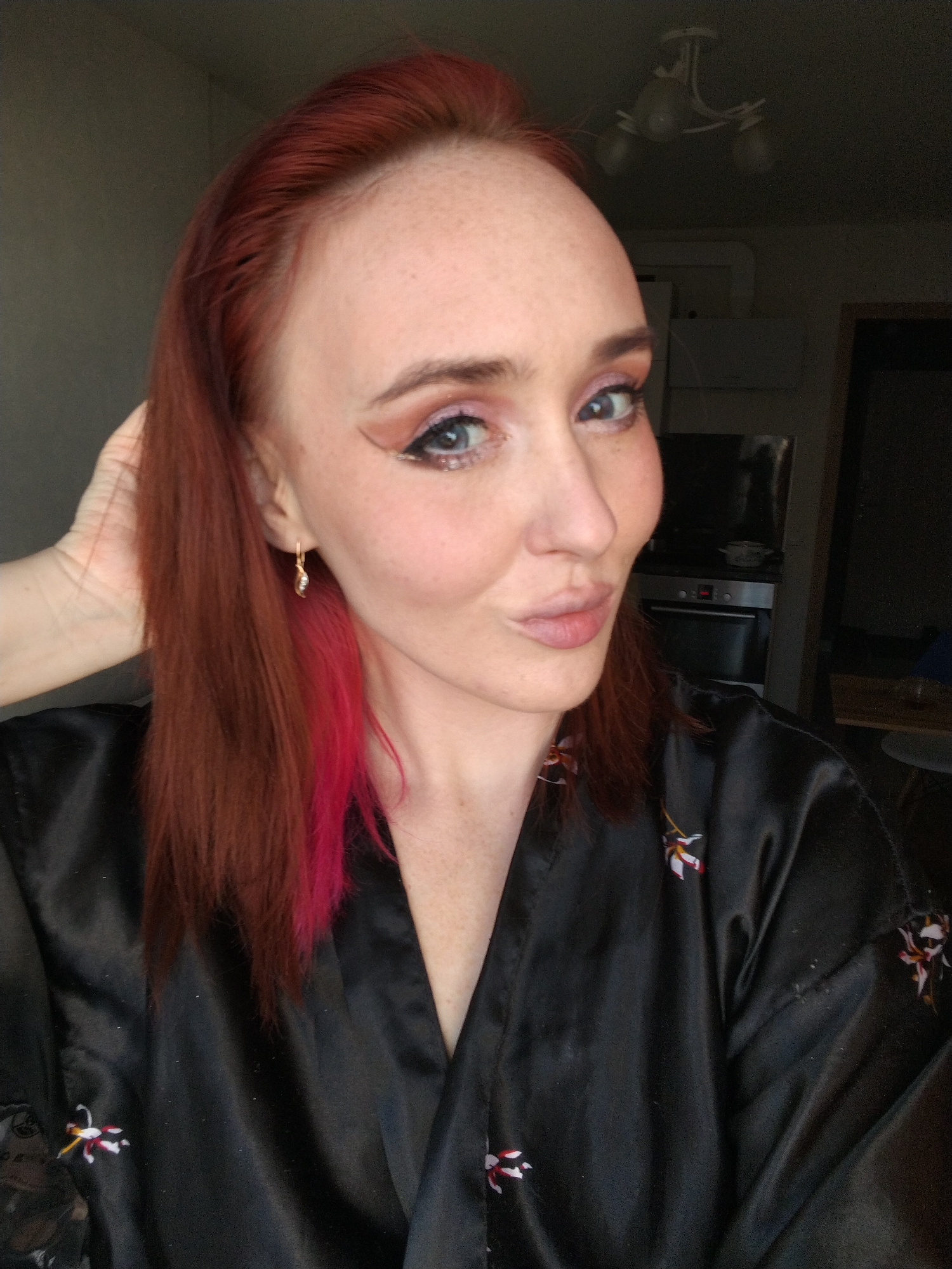 A close-up, eye-level portrait features a woman with long, reddish-brown hair, complemented by a distinct streak of bright pink hair on her right side (viewer's left). She has fair skin, blue eyes, and wears noticeable makeup, including pink eyeshadow, dark winged eyeliner, and glitter along her lower lash line. A small, gold-colored earring with a clear stone adorns her visible ear. She is dressed in a black satin-like robe or jacket, patterned with small white, red, and yellow floral designs. Her right hand is gently raised to her head, lightly touching her hair. She is looking directly at the camera with a playful, pursed-lip expression.
The background reveals a dimly lit indoor setting, likely a home environment, possibly a kitchen or living area. Partially visible elements include a white ceiling light fixture, dark kitchen appliances such as an oven and stovetop, and a dark countertop. The scene suggests an informal moment, possibly a selfie, taken indoors. No visible text is present in the image. The setting is reportedly in Izhevsk, Russia.