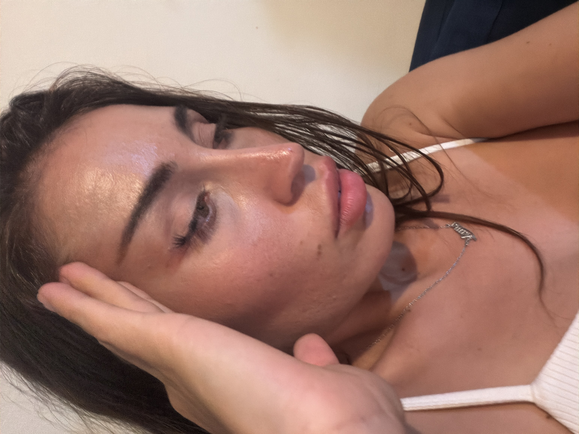 A close-up, medium shot captures a woman's face from the chest up, with her right hand held to her forehead. She has long, dark brown hair and is wearing a white ribbed top and a silver necklace with a pendant that appears to say "Mama." Her eyes are looking downwards, and her lips are slightly parted. The background is a plain, light-colored wall. The overall lighting is warm and appears to be indoor lighting. The image does not provide any contextual information about the location being Tersefanou, Cyprus.