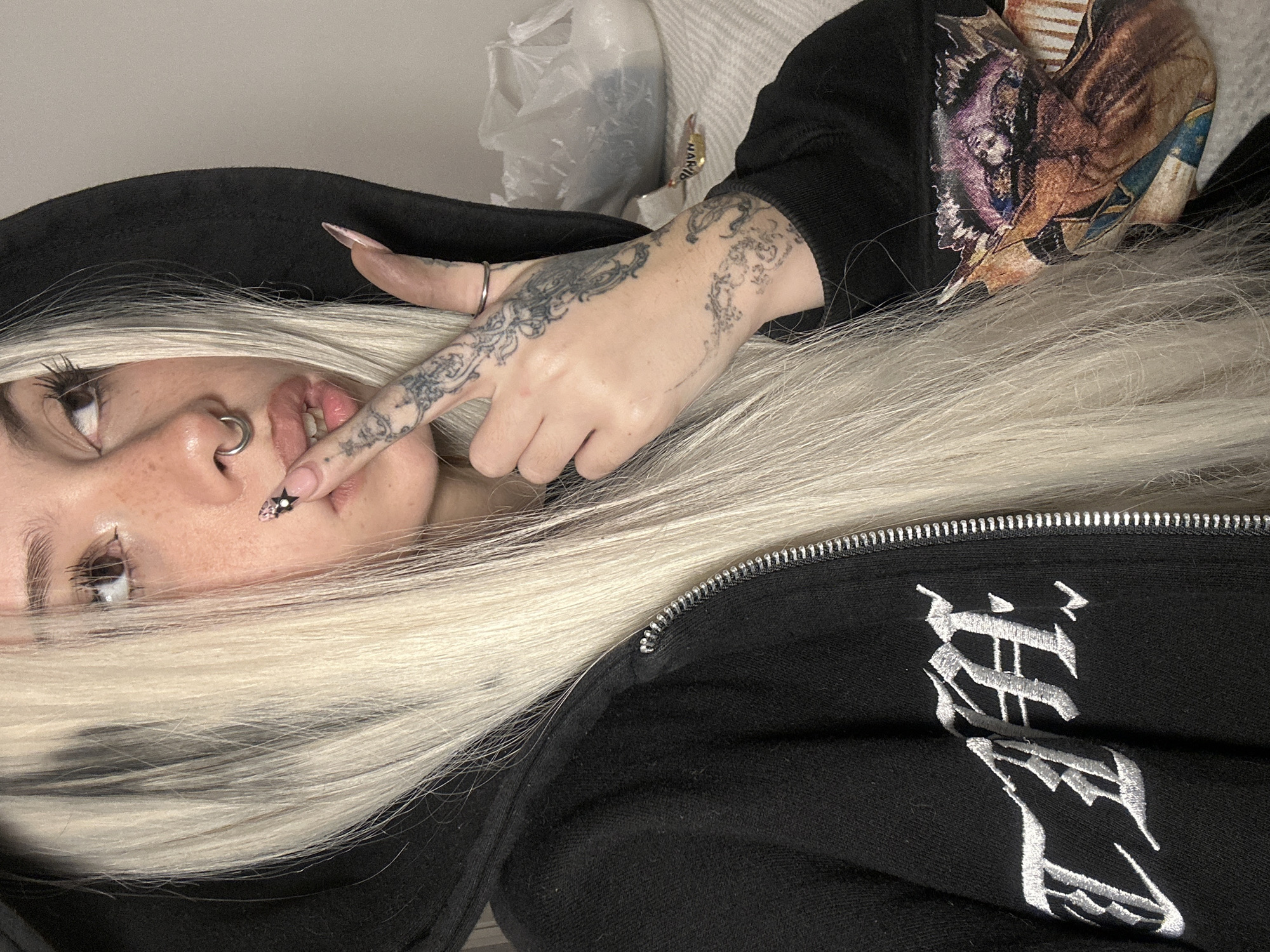 The image presents a close-up, low-angle shot of a person, likely female, with long, straight, platinum blonde hair, lying down. Their head is positioned to the left, and they appear to be looking upwards and slightly to the right. The visible features of their face include a focused eye, freckles, and a silver-colored septum piercing in the nose. Their lips are slightly parted, with a long, manicured right index finger touching the lower lip. This finger, along with the back of the hand and other fingers, is extensively covered in intricate, dark-inked ornamental or scrollwork tattoos. The index finger's nail features a distinct black star design and small embellishments, while other nails are long and a light pink/nude color. A silver ring is worn on one of the tattooed fingers.
The person is wearing a black hooded jacket with a silver zipper prominently displayed on the front. On the lower right side of the jacket, there is white embroidery in a stylized gothic or Old English script, which is partially visible. The background reveals hints of a light-colored surface, a white plastic bag, and a piece of patterned fabric, possibly a cushion or blanket, featuring an artistic design that appears to include figures or animals. The scene is set indoors, with no specific time of day or weather discernible. The pose suggests a moment of contemplation or a subtle, perhaps playful, gesture. The provided context indicates the location is Buenos Aires, Argentina, though no visual cues within the image confirm this.