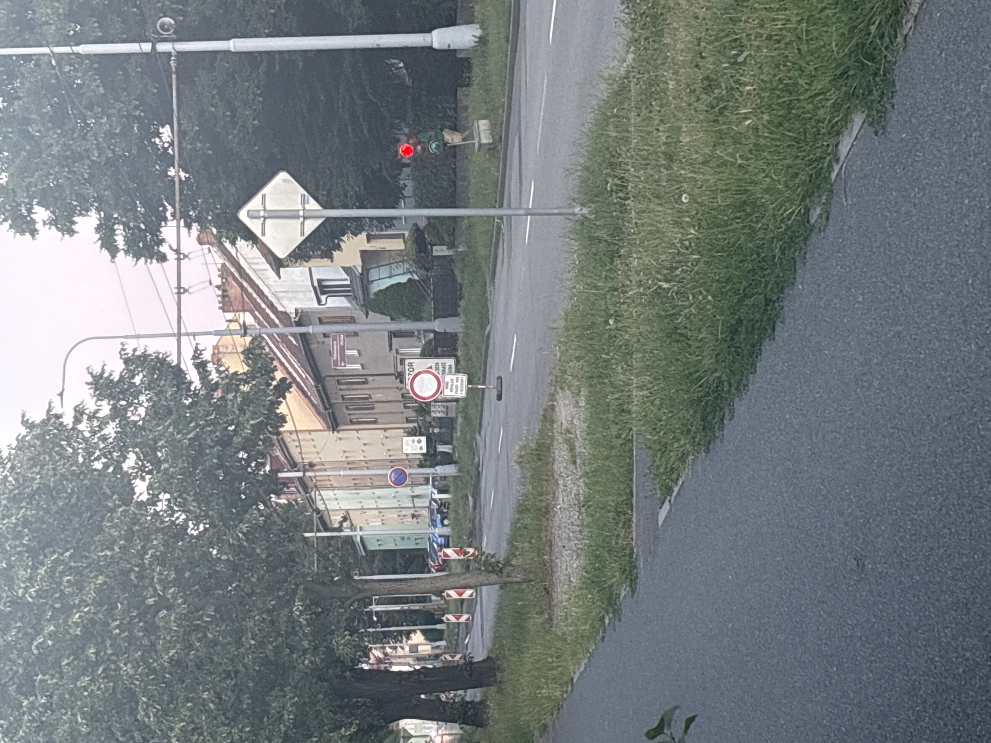 The image depicts a street scene, likely in a European town or city. The perspective is from a low angle, looking upwards and slightly to the side. The street is paved and has white lane markings. On the right side of the street, there is a strip of well-maintained green grass.
Several objects are visible. A traffic light is illuminated with a red signal, indicating a stop. A diamond-shaped traffic sign with a white background and a white cross symbol is mounted on a pole. Further down the street, there are several buildings, appearing to be houses or apartment blocks, with a mix of brick and lighter colored facades. Some of the buildings have visible windows and roof structures.
There are trees lining the street, their leaves appearing lush and green, suggesting it is likely spring or summer. The sky is visible in the upper left corner and appears overcast or very bright, with a light hue. The overall impression is of a typical urban environment during the daytime.
Visible text includes on a sign that appears to be a red circle with a diagonal line through it, and another sign with text in a European language, possibly Slovak or Czech. One sign appears to say "ZOR" followed by other text that is not fully legible. Another sign has "2016" on it. There are also smaller signs with red and white striped markings on posts along the side of the road.
The scene appears calm and orderly, with no visible people or vehicles in the immediate vicinity, apart from some parked cars obscured by trees in the distance. The lighting suggests it is daytime, possibly late afternoon or early morning given the slightly muted light.