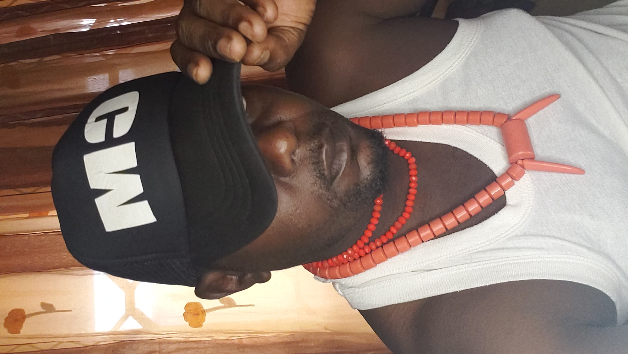 A man wearing a black trucker hat with white lettering, a white tank top, and two strands of coral-colored beads is posing for a portrait. The hat is tilted forward, obscuring part of his face, and he is holding the brim with his right hand. The beads, one thinner and one thicker with a pendant, are draped around his neck. The background consists of sheer, brown curtains with a floral pattern and a window with a white cross-bar. The lighting appears to be natural, coming from the window. The image was taken in Narayi, Nigeria.