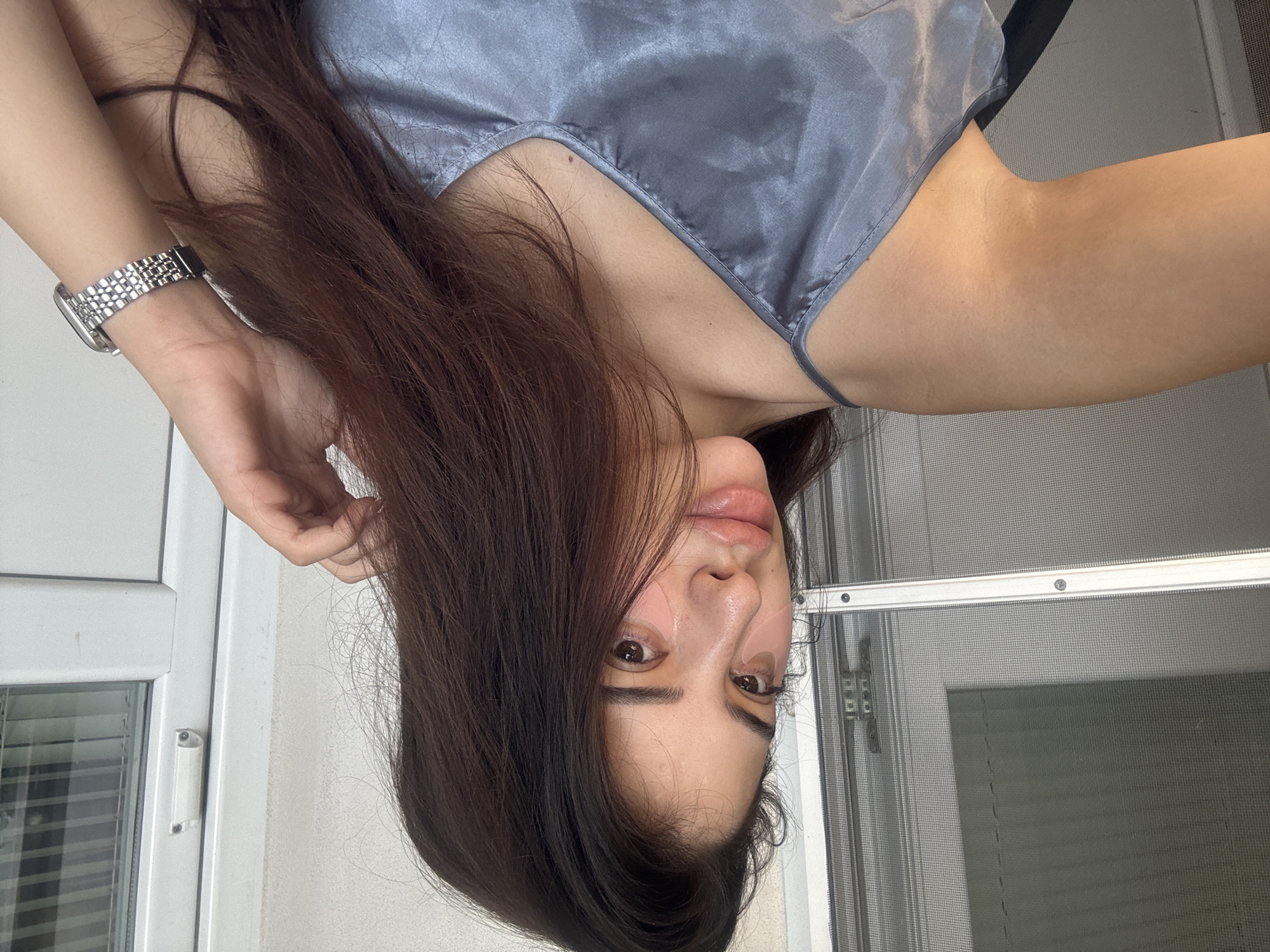 This media file captures a close-up, upside-down selfie of a person, likely female, in an indoor setting in Plovdiv, Bulgaria. The subject has long, flowing dark brown hair that cascades around their face and partially over a shoulder. They are wearing a light blue or grey camisole made of a satin-like material, featuring delicate trim along the neckline.
A metallic link bracelet watch is visible on the person's left wrist. Their face, oriented towards the bottom of the frame, shows one eye, part of the nose, and the mouth, with a neutral or gentle expression. Pink skincare patches are visible under both eyes and on the side of the nose/cheek area. A small mole is noticeable on the chest, below the camisole's neckline.
The background reveals elements of a window. On the left, a white window frame with a white latch mechanism is visible. To the right, a grey mesh screen window is prominent, with horizontal blinds partially seen through the screen, suggesting daylight outside. The image implies a personal and casual moment, possibly taken while relaxing indoors. No visible text is present.