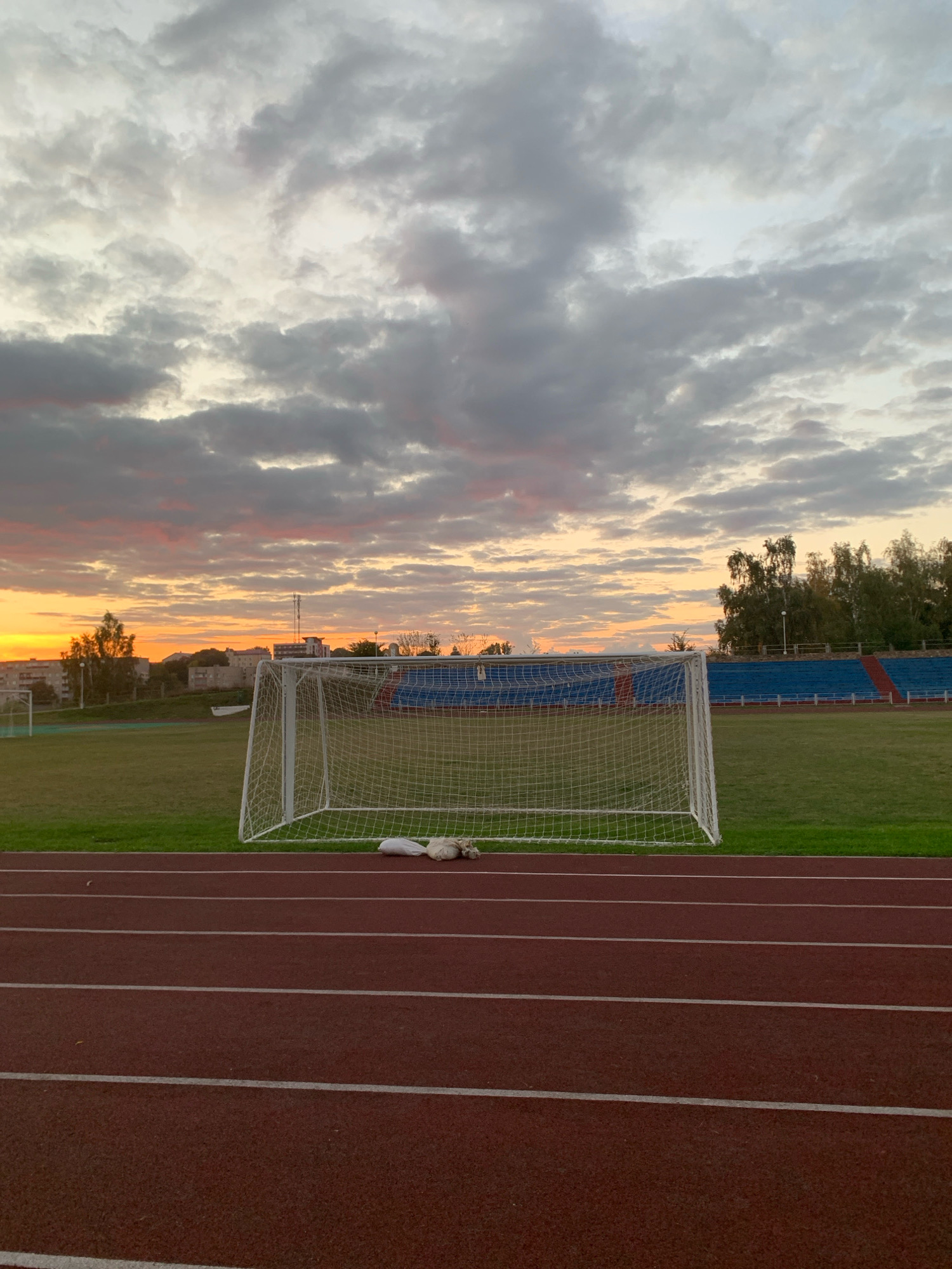 The image depicts a sports stadium in Kapyĺ, Belarus, during a sunset. The foreground features a red running track with white lines marking lanes. Beyond the track, a green field with a soccer goal is visible. The sky is filled with dramatic clouds, tinged with orange and pink hues from the setting sun. In the background, there are trees and buildings, suggesting an urban or suburban setting. A few white objects, possibly bags or clothing, are placed on the grass near the soccer goal. The overall scene evokes a sense of twilight and the end of a day.
