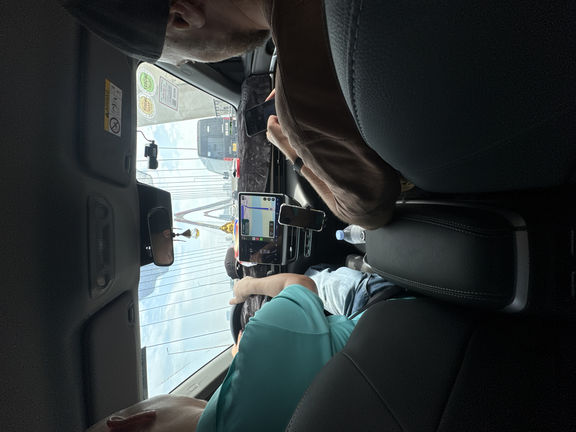 The image is taken from inside a car, likely a taxi, showing two men. The driver, wearing a turquoise shirt, is in the foreground, facing the windshield and gripping the steering wheel. A tablet device displaying a navigation app is mounted on the dashboard near the center console, along with another smartphone. A passenger is in the back seat, partially visible in the upper part of the frame, looking at his phone. Outside the car, a view of a modern suspension bridge can be seen. Based on the bridge design, the location is most likely the Nhật Tân Bridge in Hà Nội, Vietnam. A public bus can be seen directly in front of the car.