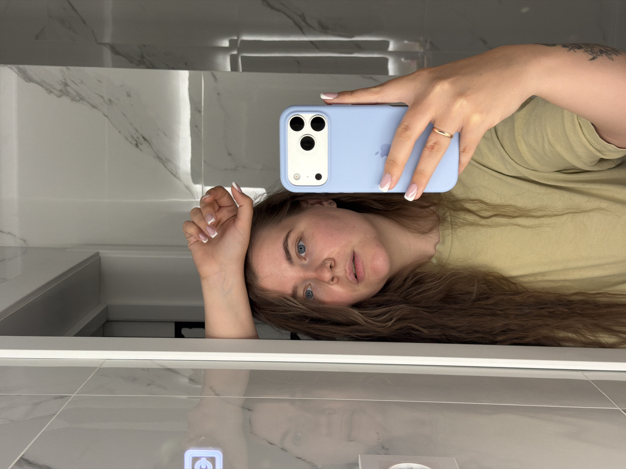 A person is reflected in a mirror, holding an iPhone 13 Pro in a light blue case. The individual has light brown hair and blue eyes, wearing a gold ring on their right hand. The background shows a bathroom with white marble-patterned tiles and built-in lighting. The location is the city of Chaikovsky, Russia.