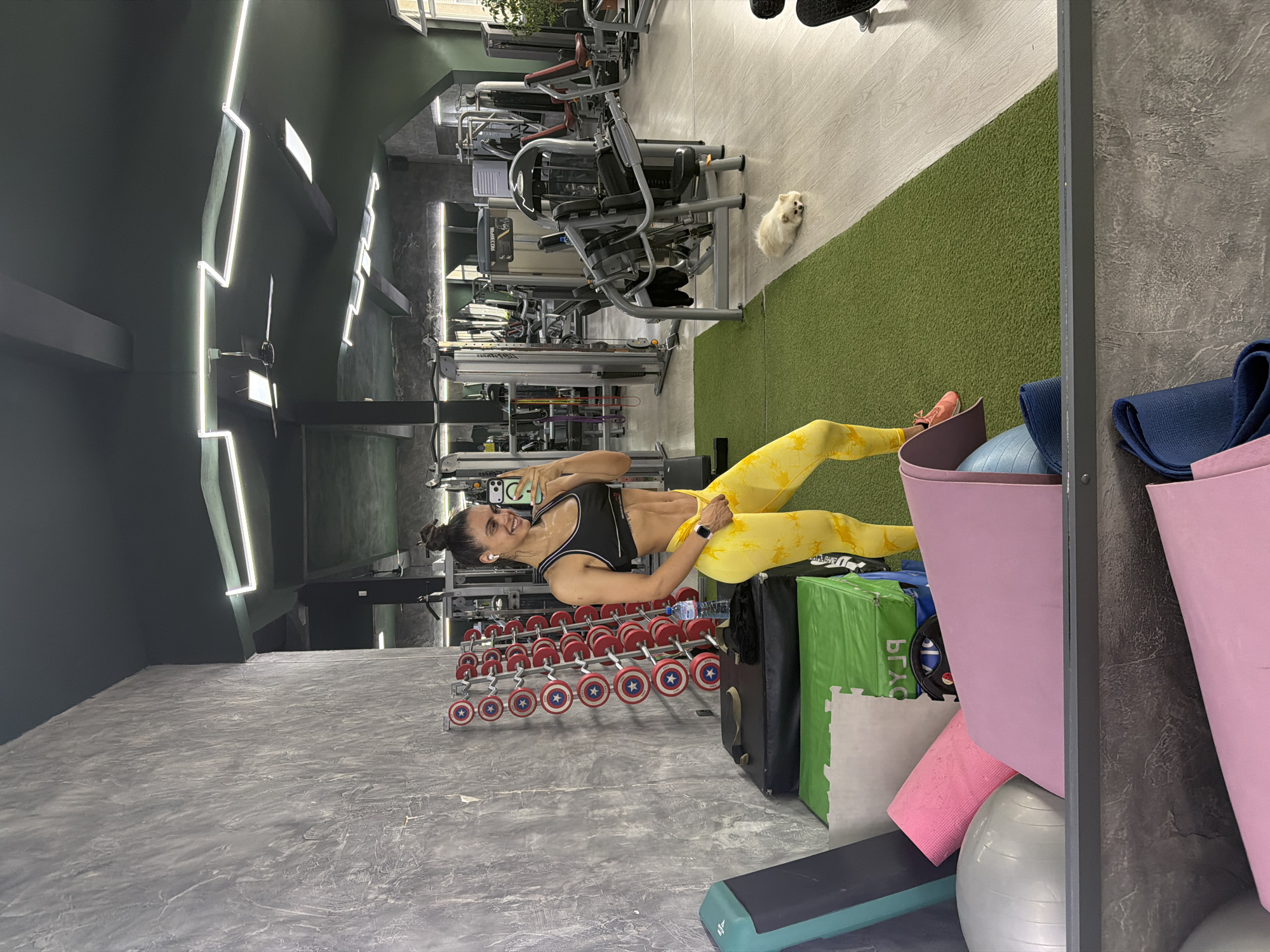A woman takes a mirror selfie in a modern indoor gym. She is in the foreground, smiling at her phone, which she holds up to capture her reflection. She is wearing a black sports bra and yellow patterned athletic leggings, with her dark hair tied back. Visible abs and a smartwatch on her wrist suggest a fit physique.
Behind her, an array of red dumbbells with blue and white Captain America shield designs are neatly arranged on a rack. A water bottle rests on top of this rack. In the mid-ground, a small white fluffy dog lies calmly on a green artificial turf section of the floor. Beyond the dog, various workout machines, including weight machines, are visible.
The gym features contemporary design elements, with dark green and textured grey concrete walls. The ceiling and walls are adorned with bright, linear LED light fixtures, providing ample illumination. The floor is a mix of the green turf, light-colored wooden or laminate flooring, and the grey concrete texture.
To the right of the woman, various gym accessories are stored: rolled-up blue and pink yoga mats, a grey exercise ball, and pink storage bins. A green bag or box, partially obscured, shows some reversed text, possibly "PRAX" or "PRAG." A workout step is also partially visible on the floor. The overall atmosphere is clean and well-lit, suggesting an active and well-maintained fitness environment.