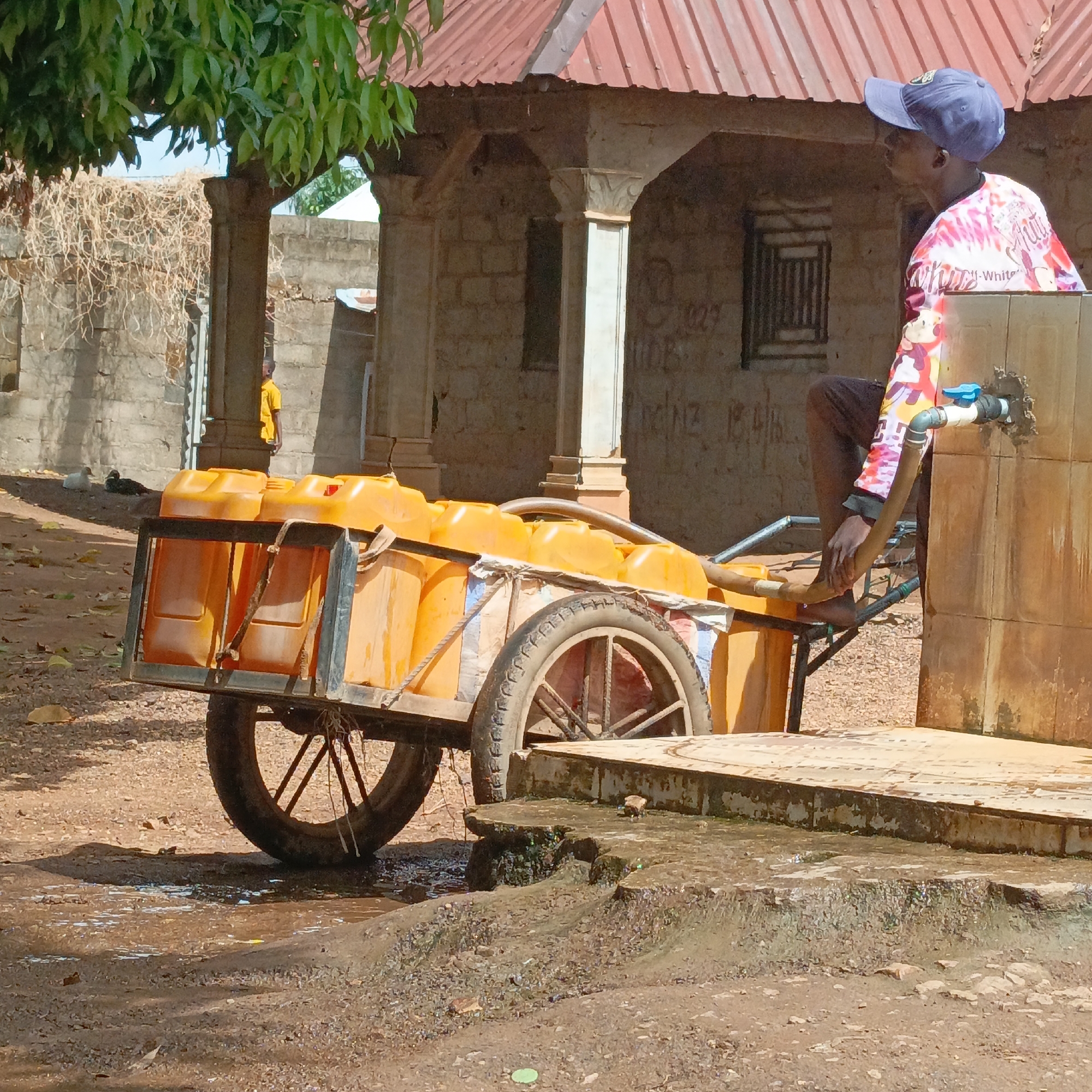 In Akwanga, Nigeria, a man in a blue cap and colorful shirt sits on a cart laden with orange jerrycans. He appears to be filling the containers with water from an outdoor tap attached to a tiled wall. The cart has two wheels with visible spokes. A building with a red metal roof and concrete pillars is in the background, along with another structure partially visible. The ground around the tap is wet, suggesting recent use.