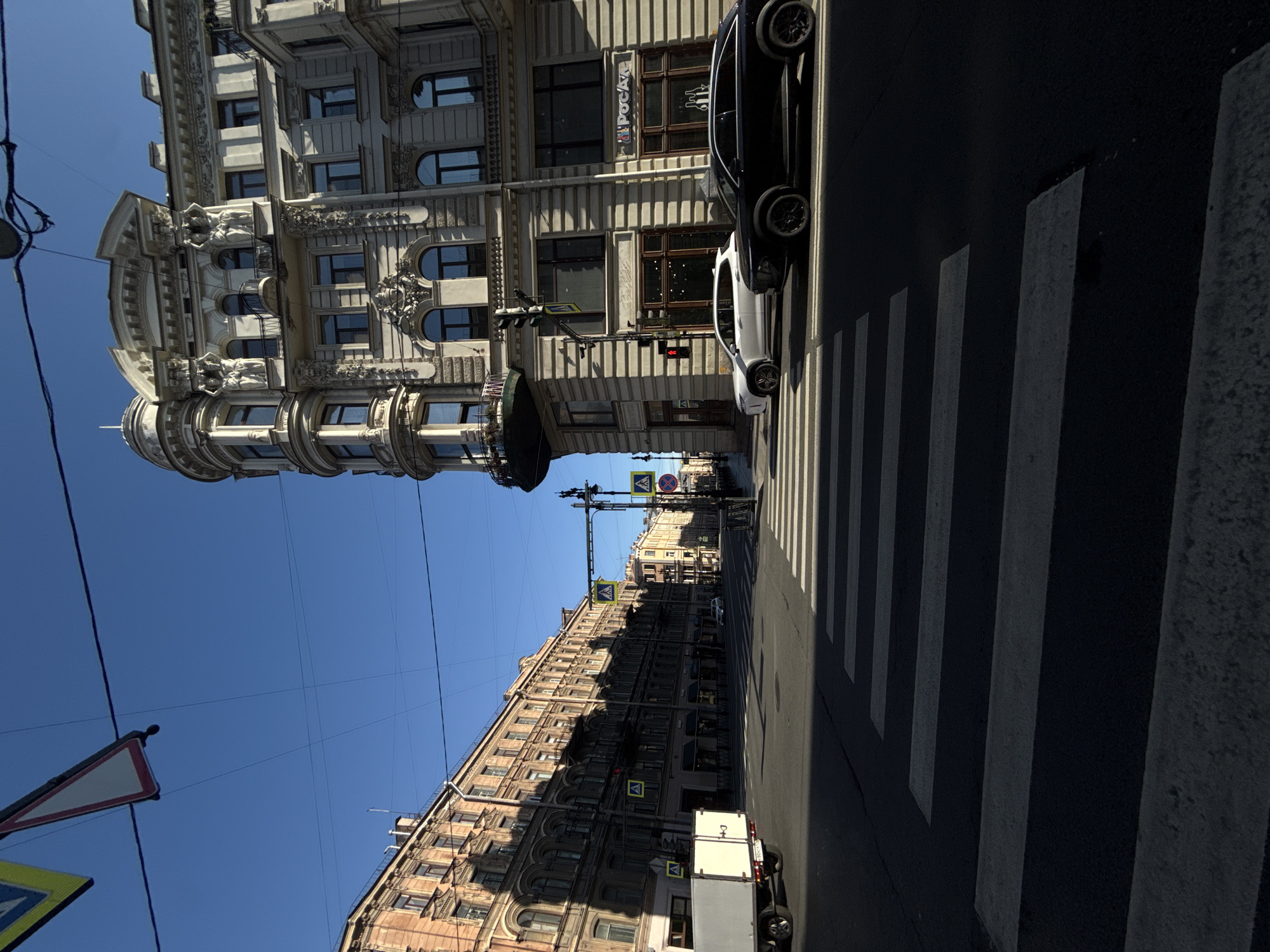 The image captures an urban street scene in Saint Petersburg, Russia, with a clear blue sky overhead. A pedestrian crossing is prominently featured in the foreground, with stark white stripes contrasting against the dark asphalt. Two cars, a white SUV and a black SUV, are positioned at the crossing.
To the right, an ornate, multi-story building with classical architectural details stands tall. Sculptural figures adorn its facade, and balconies project from some of its windows. The building's facade is light-colored and features rows of windows. A sign with Cyrillic lettering is visible on the ground floor.
To the left and extending into the distance, a long, multi-story building with a reddish-brown facade lines the street. This building also has multiple windows and appears to be older in style. A white truck with a closed trailer is parked on the left side of the street, partially in shadow.
Traffic lights and pedestrian crossing signs are visible at the intersection, indicating a regulated street crossing. Overhead wires crisscross the sky, a common sight in urban environments. The overall scene suggests a bright, sunny day in a European city.