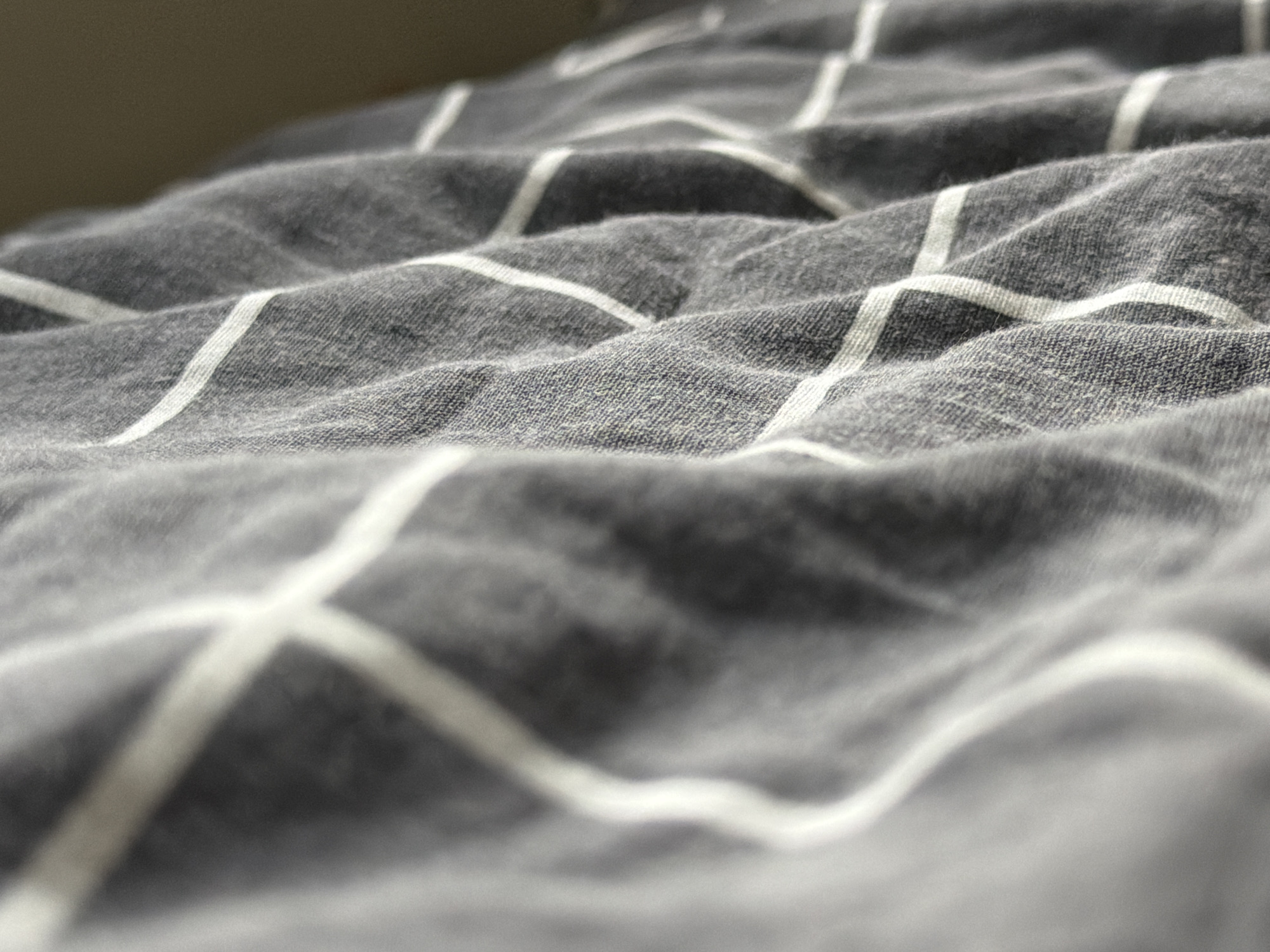 The image displays a close-up, shallow depth-of-field view of a wrinkled gray fabric with a white striped pattern. The stripes form a grid-like design, and the fabric appears to be a bedsheet or similar textile. The lighting suggests an indoor setting, possibly a bedroom. The background is out of focus and appears to be a plain, neutral color, possibly a wall or a piece of furniture. There are no people, animals, or any other discernible objects or actions in the image. The scene is static and focuses entirely on the texture and pattern of the fabric. There is no visible indication of any location in the city of Khimki, Russia, or any urban environment.