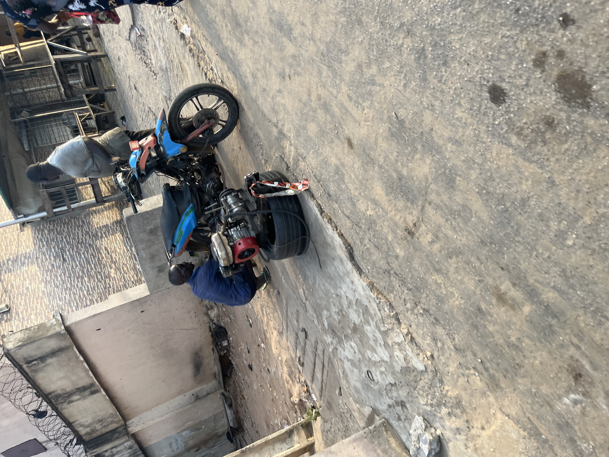 In an outdoor urban environment, a man in a blue shirt and sandals is crouched down, working on the engine of a blue motorcycle. The motorcycle is parked on a concrete surface with some debris, including a detached tire and engine parts, scattered around it. In the background, another man wearing a light-colored shirt and apron stands near a wooden structure. A building with a textured facade and barbed wire on top is visible to the left. The scene is illuminated by natural sunlight, casting shadows on the ground.