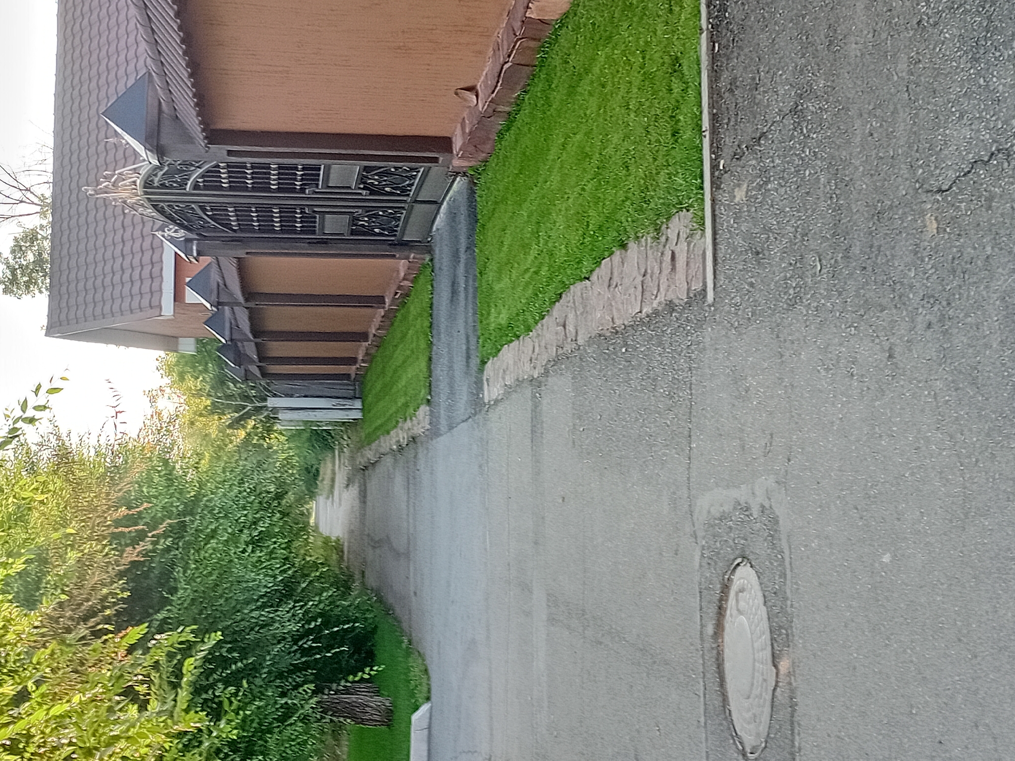 This is a photograph of a paved pathway in Semey, Kazakhstan, bordered on the left by a line of trees and on the right by a decorative metal gate and a stuccoed wall. The pathway appears to be a sidewalk or a private driveway. The gate is ornate, featuring intricate scrollwork and finials, and is set within a substantial wall. The wall is a uniform reddish-brown color and is topped with decorative caps on its posts. In front of the wall and gate, a neatly manicured strip of bright green grass is visible. The pathway itself is asphalt, showing some signs of wear and tear with visible cracks and seams. A circular manhole cover is embedded in the pathway towards the bottom left of the image. The lighting suggests it is daytime, possibly late afternoon, as the sun casts a warm glow on the scene. The overall impression is one of a well-maintained, perhaps residential, area with a focus on both security and aesthetics.