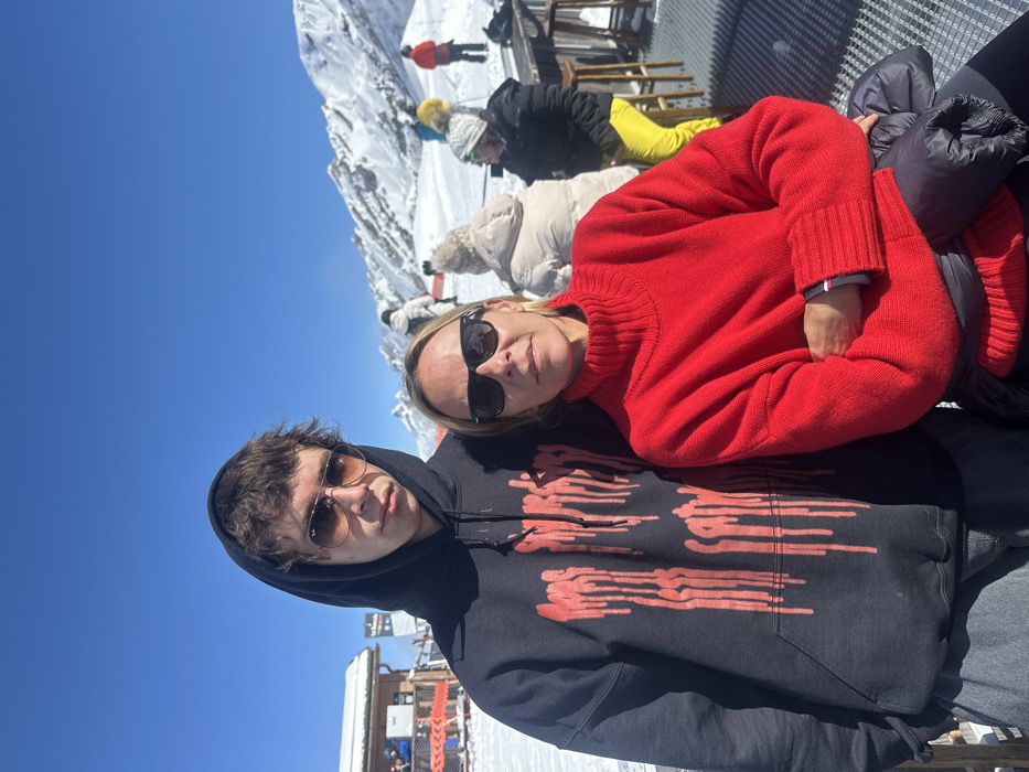 The image depicts two individuals posing for a photograph on a sunny winter day at a ski resort in La Clusaz, France. The foreground features a young male on the left and an adult female on the right. The male has light brown hair, wears dark aviator-style sunglasses, a black hooded sweatshirt with a red "dripping" graphic design, and dark trousers. He stands with a neutral expression, looking directly at the camera. The female wears dark rectangular sunglasses, a bright red ribbed turtleneck sweater, and has blonde hair. Her arms are folded across her stomach, with a black puffer jacket resting on her lap, and she also looks directly at the camera with a neutral expression. They are positioned on a dark gray metal grated surface, possibly an outdoor patio.
In the midground and background, several other people are visible, dressed in winter ski attire, some seated at wooden bar-height tables and stools on the snowy ground. To the far left, a multi-level wooden building, typical of a mountain lodge or restaurant, is partially visible. The background is dominated by extensive snow-covered terrain and prominent snow-capped mountains under a clear, brilliant blue sky, indicative of alpine conditions. The scene is brightly illuminated by natural sunlight, with crisp shadows suggesting a high sun angle.