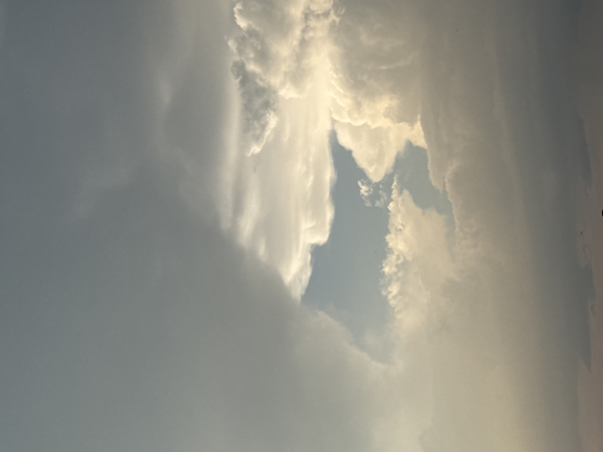 This media file captures a dramatic sky with thick, textured clouds. The clouds are varied in color, ranging from dark gray and muted tones on the left to bright, luminous whites and yellows on the right, suggesting sunlight breaking through or reflecting off their surfaces. A large gap in the clouds reveals a patch of light blue sky. The composition is vertical, as if the camera was pointed upwards. The lighting implies it is either early morning or late afternoon, with the sun low on the horizon. The overall mood is one of atmospheric intensity and natural beauty. There is no visible text.