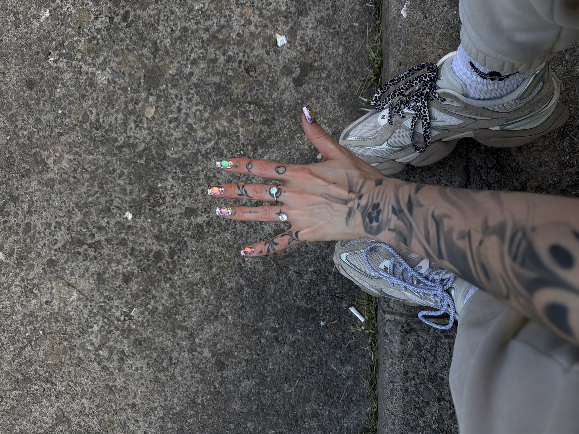 An overhead, close-up shot captures a person's tattooed arm, hand, and feet on a concrete surface, likely a sidewalk or street in Budva, Montenegro. The person is wearing light-colored (beige/grey) joggers and light-toned New Balance sneakers. One shoe features distinct black and white cheetah print laces, while the other has light blue laces. White socks with a black embroidered design (possibly a stylized wave or smile) are visible above the sneakers.
The prominent left hand is heavily adorned. The fingers display a variety of press-on or artificial nails with an iridescent, gem-like finish, showing colors like green, orange, pink, and purple. Several rings are worn on the fingers, including one with an opal-like stone, one plain band, and one with a large clear stone. Small, simple tattoos (stick-and-poke style) are visible on the fingers and knuckles, including a rose, geometric shapes, and what appears to be an alien head. The arm is covered with a full sleeve tattoo featuring intricate black and grey designs.
The concrete ground is rough and varied in texture, with small pebbles and bits of debris scattered across it, including a cigarette butt near the right foot and a piece of crumpled paper or plastic further up. Hints of dry grass are visible along the edge of the concrete, suggesting a curb or border. The lighting is soft and even, indicating daytime, possibly overcast or shaded, as there are no harsh shadows. The overall scene provides a detailed glimpse into the person's personal style and immediate surroundings. The "NB" logo is visible on the sneakers, signifying the New Balance brand.