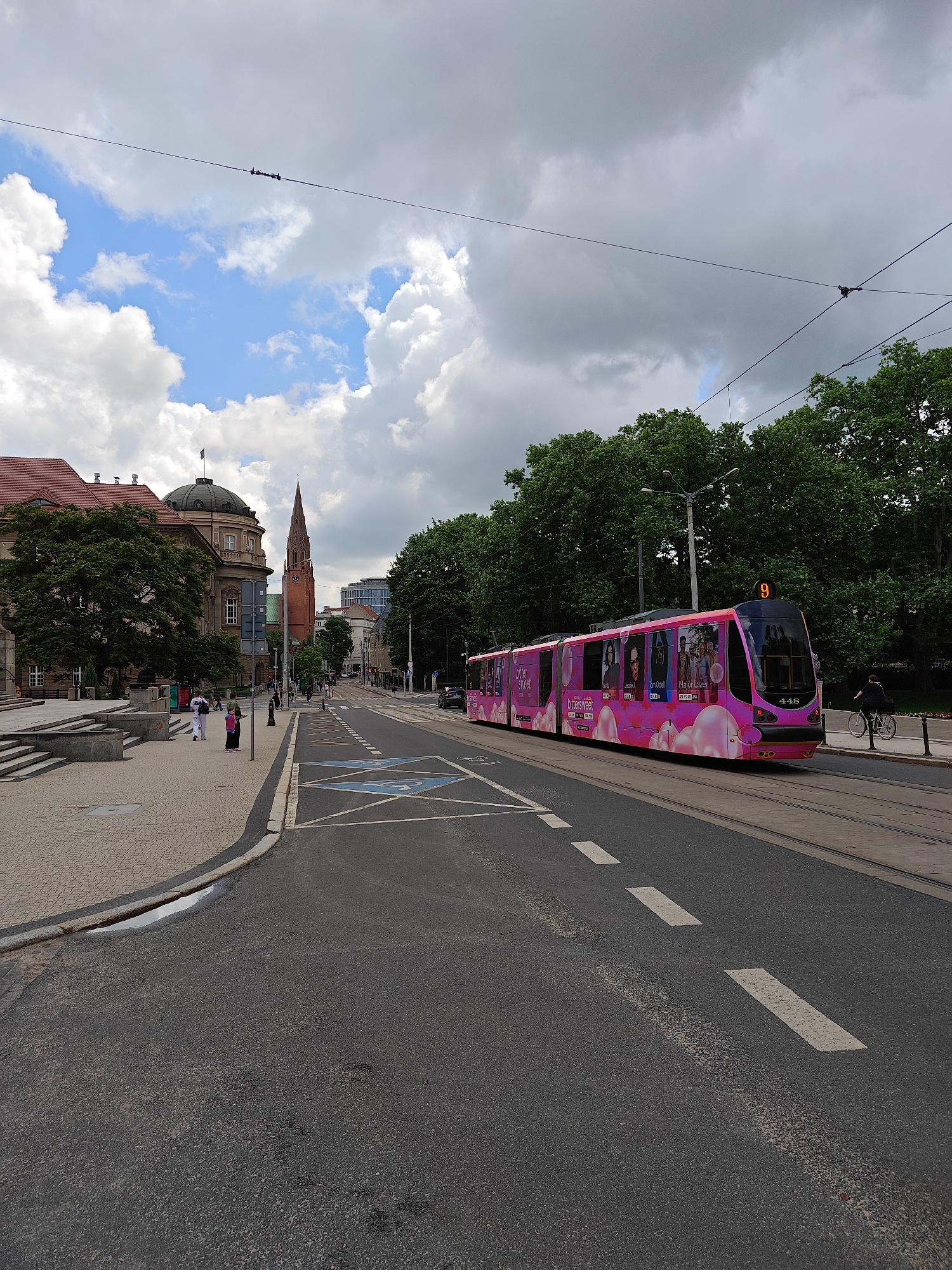 A pink tram with advertisements is moving along a street in a city. The tram is decorated with images of people and the word "bittersweet". It has the number 9 displayed on its front. A person on a bicycle is visible on the right side of the tram. In the background, there are trees and buildings, including a church with a tall spire and a building with a dome. The street has tram tracks and road markings. The sky is cloudy with patches of blue. There are a few pedestrians on the sidewalk on the left side of the street.