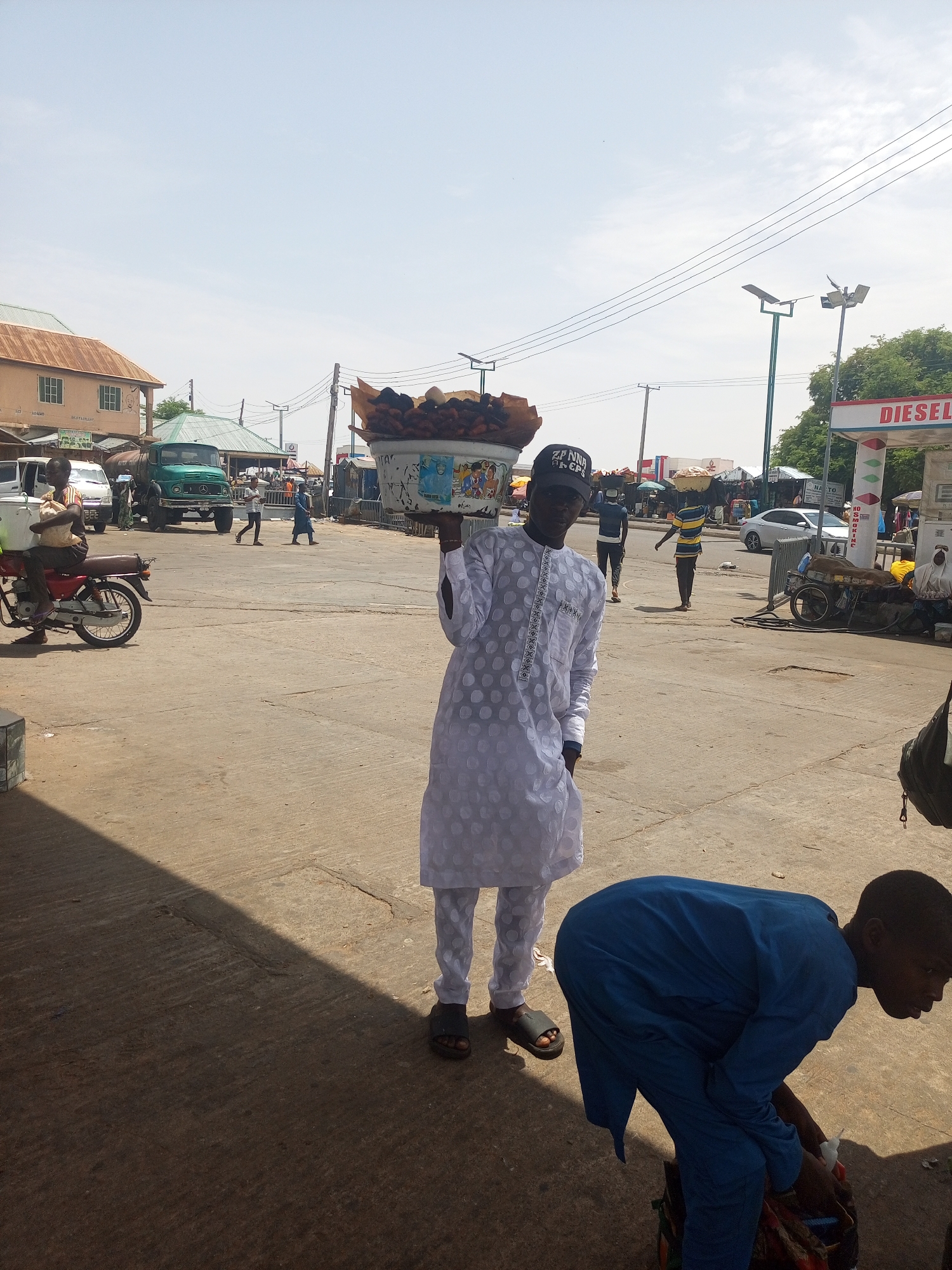 An eye-level medium shot depicts a street scene in Gombe, Nigeria. A man dressed in a white, patterned tunic and matching pants and a black baseball cap holds a large basket filled with what appears to be cooked food items on his head. A child in a blue outfit bends over near the lower right corner. In the background, other individuals, vehicles, and buildings are visible, including a green truck and a gas station. The sky is lightly clouded.
