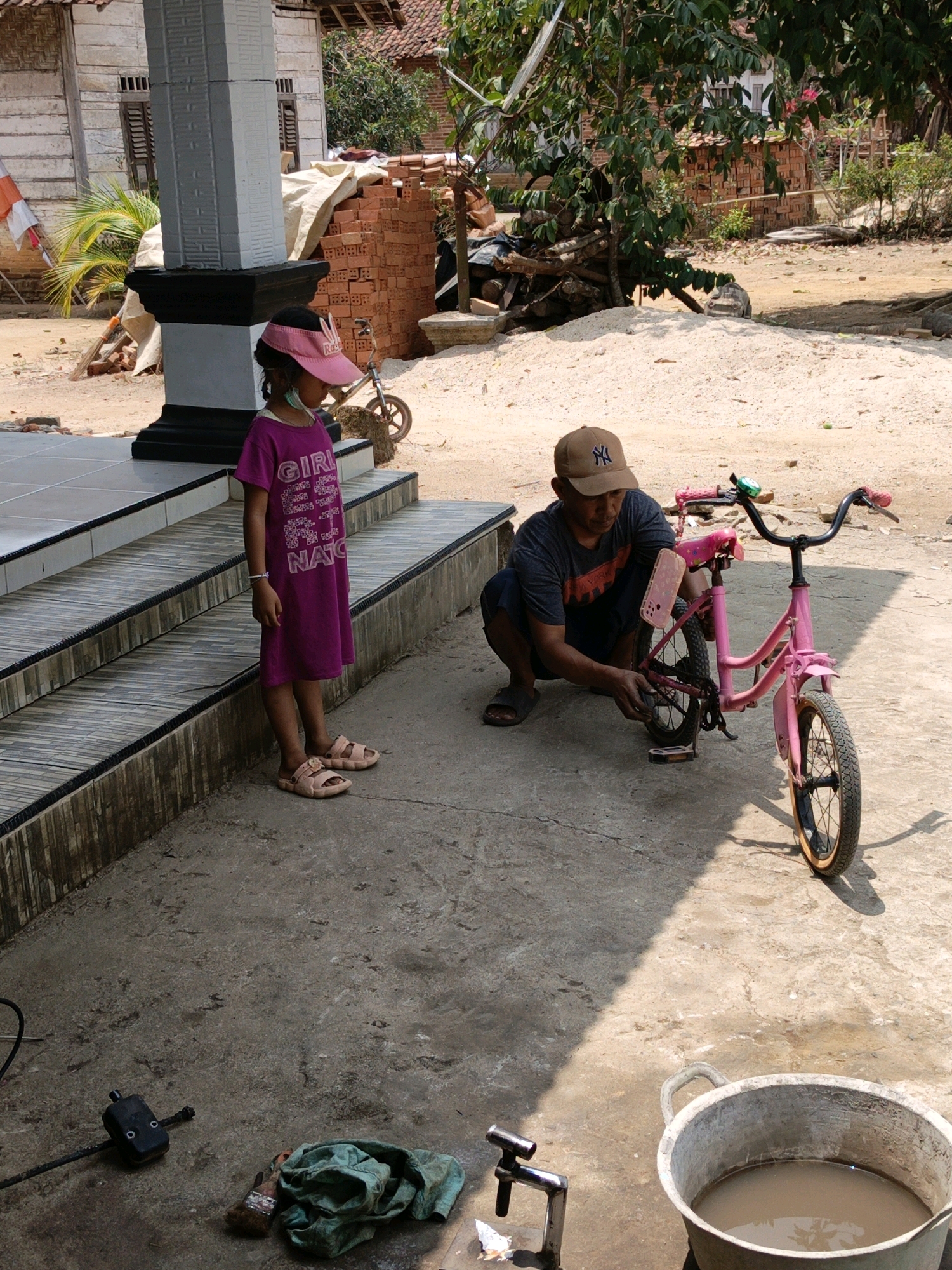 In Gedung Nyapah, Indonesia, a man kneels on a concrete ground, tinkering with a pink bicycle. He wears a brown baseball cap and a dark t-shirt. Beside him, a young girl stands on a tiled staircase, looking down. She has dark hair and wears a pink cap and a purple dress. The scene is outdoors with a building and scattered bricks in the background. A large metal bucket filled with murky water sits on the ground near a faucet and some green cloth.