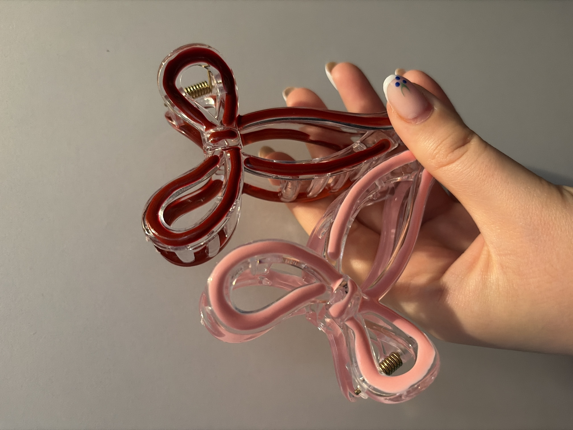 A close-up image depicts a fair-skinned hand, likely female, holding two bow-shaped hair claw clips against a plain, light gray background. The hand is positioned centrally, angled slightly from the lower right, with fingers gripping the clips.
The left hair clip is transparent plastic with a light pink inner structure forming the bow loops and center knot. A gold-colored coiled spring is visible within its hinge mechanism on the left side. The right hair clip is also transparent plastic, featuring a dark red inner structure similarly forming a bow. A corresponding gold-colored coiled spring is visible within its hinge mechanism on the right side. Both clips appear to be open or slightly ajar, showcasing their design.
The hand exhibits well-groomed fingernails. The visible thumbnail, facing the viewer, features a French manicure with a white tip, adorned with three small blue dots arranged in a triangular pattern near the cuticle. The other visible fingernails also appear to have white tips.
The background is uniformly plain, light gray, and devoid of any identifiable features, textures, or objects, offering no contextual information regarding the environment or specific location. There are no other individuals present in the frame. The image does not contain any visual data to ascertain its geographical location, including the city of Dobrich, Bulgaria.