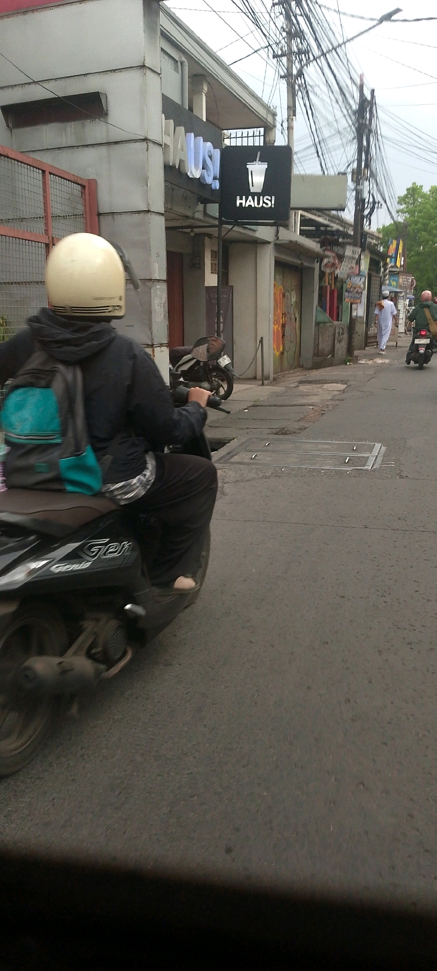 In the city of Bandung, Indonesia, a person wearing a cream-colored helmet and a dark hooded jacket with a teal and gray backpack is riding a black motorcycle. The person is wearing black pants and appears to be barefoot, with one foot visible on the motorcycle's footrest. The motorcycle has the word "Gen" and "Genio" visible on its side. The rider is moving on an asphalt road with visible manhole covers. To the left of the road, there is a building with a sign that reads "HAUS!" in large, illuminated blue letters, and a smaller black sign with a white cup icon and the text "HAUS!". The building's facade is made of light-colored metal panels. Further down the street, another person wearing a white robe walks away from the camera, and a second motorcycle with a rider wearing a green jacket and a pink helmet is also visible. The environment appears to be a commercial street with utility poles and wires overhead, and trees visible in the background. The sky is overcast.