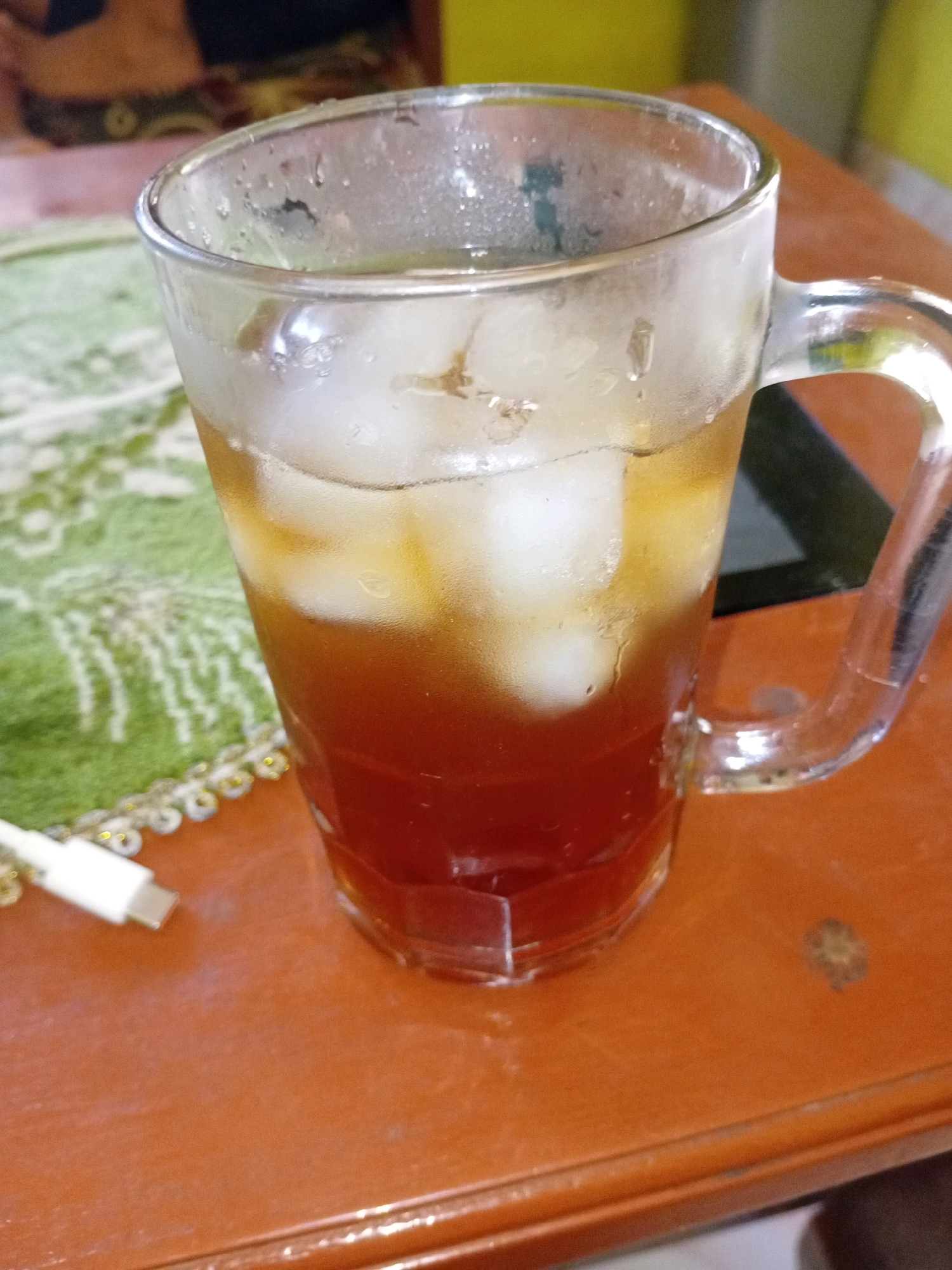 A close-up shot shows a clear glass of iced tea sitting on a brown wooden table. The tea is dark amber in color, with ice cubes visible throughout. The glass is sweating, indicating it is cold. To the left of the glass is a white USB cable. The table is set on a green mat with an intricate white pattern. In the background, a blurry image of what appears to be people and a yellow wall can be seen. The overall impression is a casual, indoor setting, likely a home or cafe, possibly in the city of Aditirto, Indonesia, given the context provided.