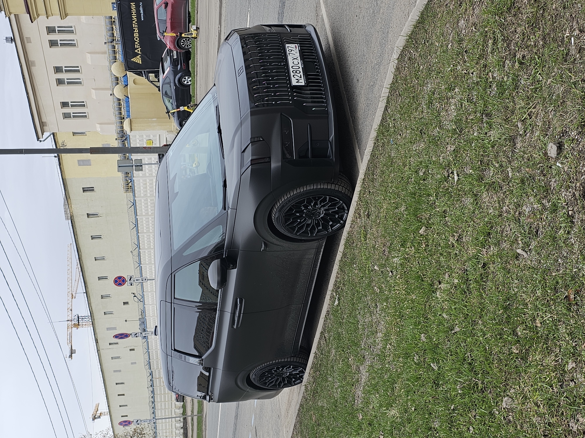 A matte black minivan is parked on the side of a street in Khimki, Russia. The vehicle has a distinctive front grille and black rims. The license plate is visible and reads "M280CX 797 RUS". The vehicle is parked next to a curb and a grassy area. In the background, there is a tall building with multiple windows, traffic signs, a construction crane, other cars, and a black delivery truck. The sky is overcast.