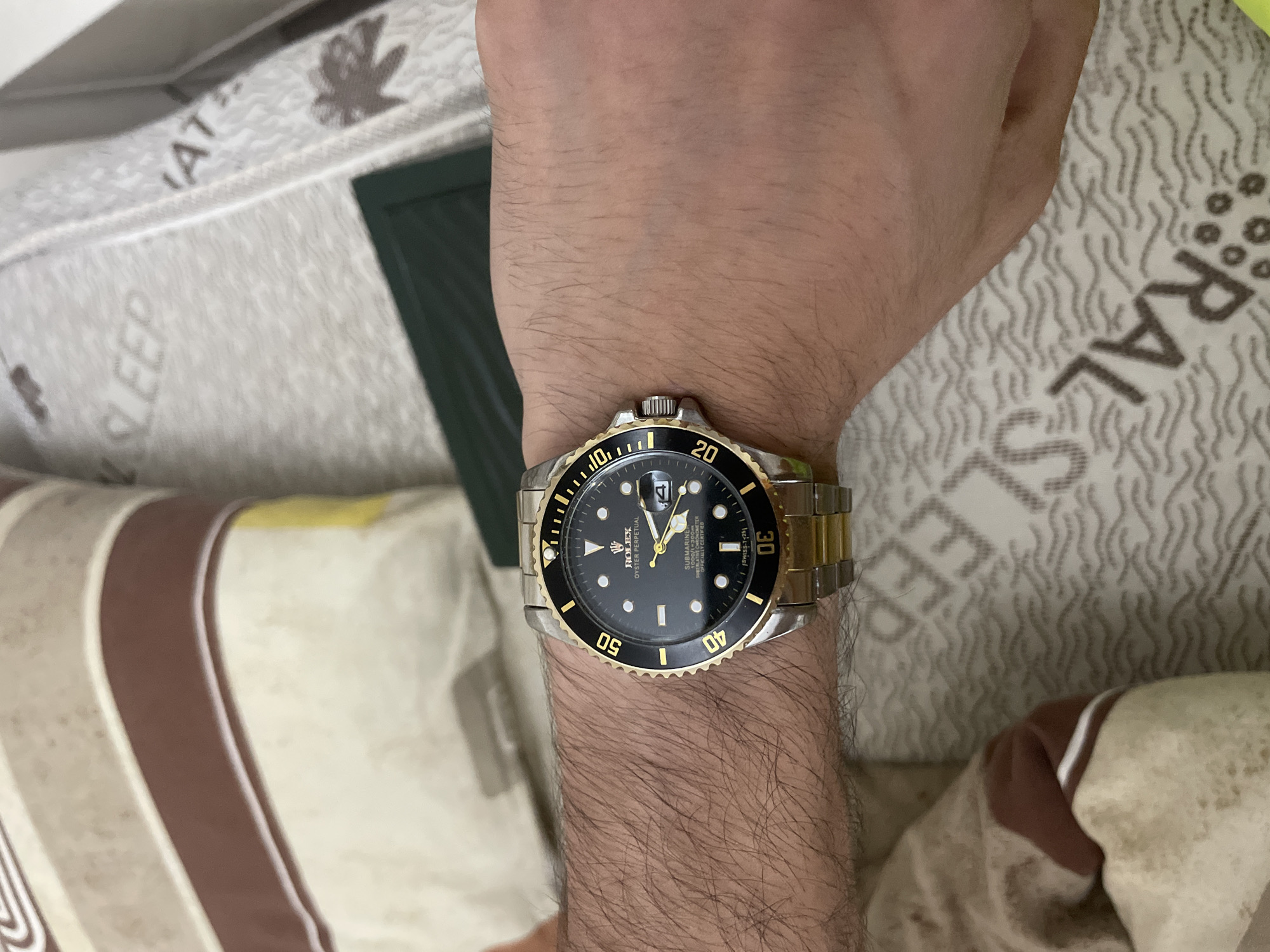 A close-up shot captured in Ihtiman, Bulgaria, shows a person's hairy arm and hand, prominently featuring a watch on the wrist, against a background of patterned bedding. The subject is likely indoors, possibly in a bedroom, and appears to be resting or lying down.
The main subject is a two-tone Rolex Oyster Perpetual Submariner watch. It features a silver and gold bracelet, a gold fluted bezel with a black insert, and a black dial. Notable details on the watch face include the brand "ROLEX" and model names "OYSTER PERPETUAL" and "SUBMARINER." Other text elements are "1000ft=300m," "SUPERLATIVE CHRONOMETER OFFICIALLY CERTIFIED," and "SWISS <T> 25." The bezel displays gold numerals at the 10, 20, 30, 40, and 50-minute marks, and the watch has a date window with a cyclops magnification lens.
The background consists of light-colored bedding, likely a mattress cover or pillow, adorned with a wavy pattern and scattered text. The word "SLEEP" is clearly visible multiple times, along with partial words such as "RAL" (possibly from "NATURAL") and "AT." A dark green rectangular object is partially visible behind the bedding. The overall impression is a casual, personal shot focusing on the watch.