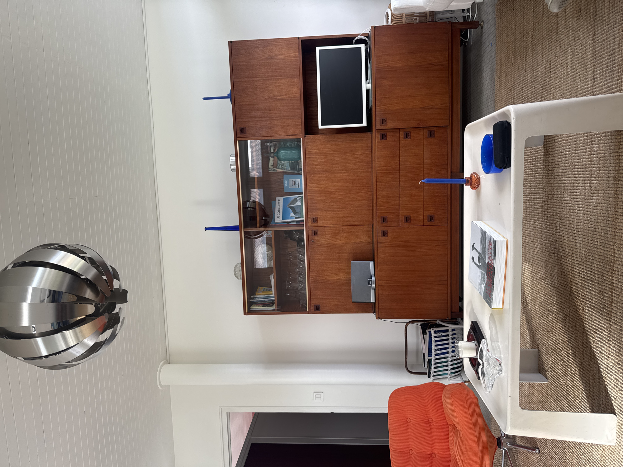 A room in Soulac-sur-Mer, France features a vintage mid-century modern design. A large teak wall unit dominates the wall, incorporating a built-in television, a glass-fronted display cabinet, and multiple storage compartments. The wall unit is adorned with blue decorative objects. In the foreground, a white lacquered coffee table holds books, decorative glassware, and a blue container. An orange office chair is positioned near the coffee table. A modern chrome pendant light hangs from the white, paneled ceiling. A doorway is visible in the background, leading to another room with purple walls. The floor is covered with a beige rug.