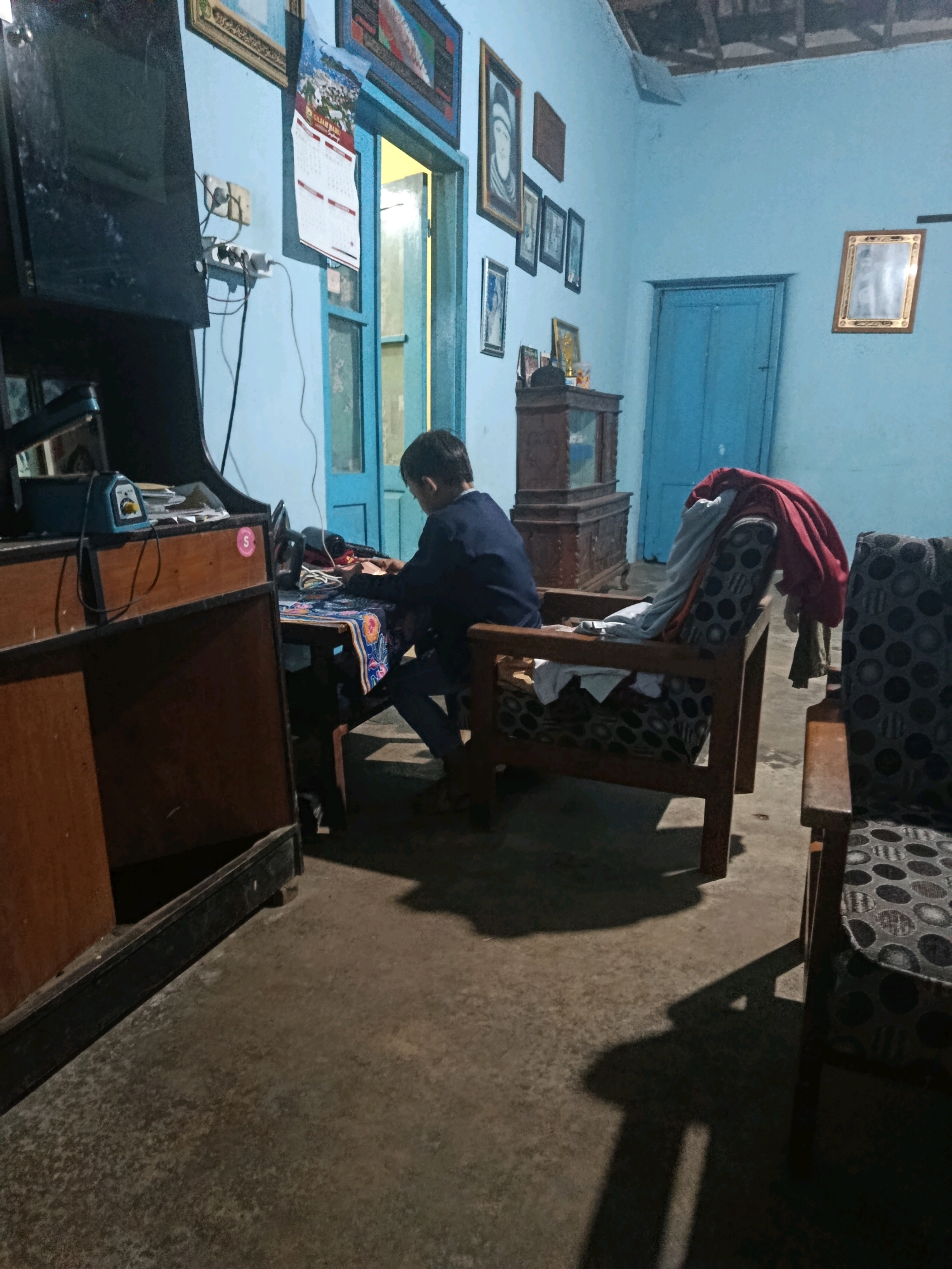 This is a forensic, matter-of-fact description of an indoor scene in the city of Sepanjang, Indonesia. The image depicts a dimly lit room, likely a living area or study, with a young person seated at a table.
**Environment:** The room has light blue walls and a visible ceiling with exposed wooden beams. The floor appears to be concrete. There are several framed pictures on the walls, including what looks like a calendar. Two wooden doors are visible, one slightly ajar, revealing a brighter interior beyond. Furniture includes a wooden desk or cabinet on the left, a wooden table with a patterned cloth in the center where the person is working, and two upholstered armchairs on the right, one of which has several garments draped over it. A dark, flat-screen television or monitor is mounted on the left side of the frame. Electrical outlets and cords are visible on the wall.
**People and Actions:** A single individual, appearing to be a young person with dark hair, is seated at the table in the center of the room. They are wearing a dark blue long-sleeved shirt and dark trousers. The individual is leaning forward, with their hands positioned over the table, suggesting they are engaged in an activity such as writing, drawing, or using a device. Their gaze is directed downwards at the table.
**Objects:** Key objects include the television/monitor, a desk/cabinet, a table with a patterned tablecloth, a chair the person is sitting on, two armchairs with patterned upholstery, framed pictures and a calendar on the walls, and various pieces of clothing. Electrical appliances and cords are also present.
**Lighting and Atmosphere:** The lighting is low, casting shadows across the floor. The light source appears to be coming from the open doorway and potentially from the activity the person is engaged in, though no direct light source is clearly visible on the person or table. The overall atmosphere is subdued and appears to be nighttime or a very dark day.