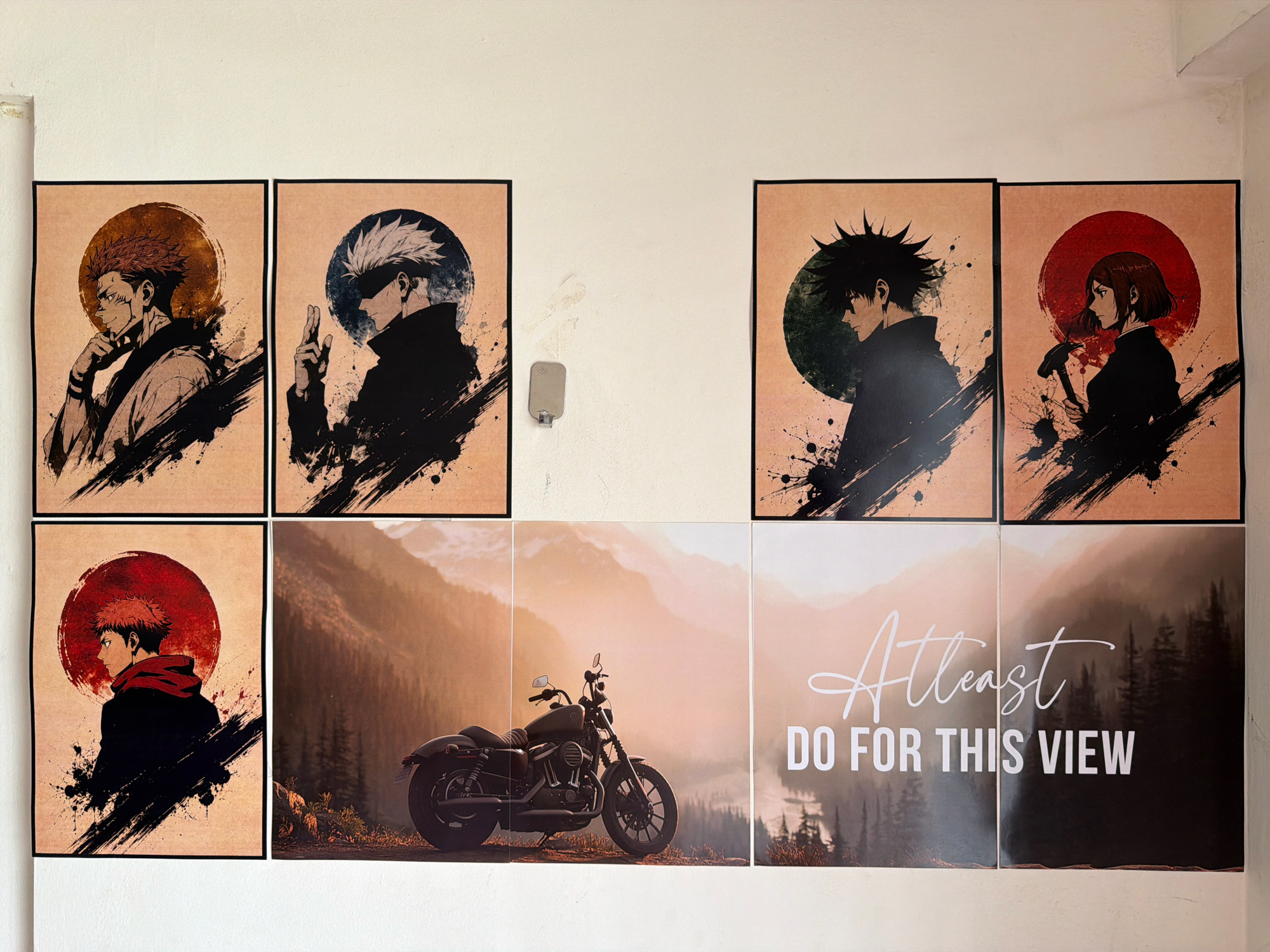 The image depicts a wall adorned with several framed posters. The top row features four anime-style illustrations, each with a circular background element. From left to right, the first poster shows a character with spiky red hair and a thoughtful expression, the second a character with white hair and a blindfold, the third a character with spiky black hair in profile, and the fourth a character with short brown hair holding a hammer. The bottom row consists of three landscape-oriented posters. The leftmost poster shows a character with spiky red hair and a red scarf in profile, similar to the top-left poster. The middle and rightmost posters are a diptych of a motorcycle parked on a scenic overlook with mountains and trees in the background. The rightmost poster of the diptych also contains white text that reads "Atleast DO FOR THIS VIEW". The overall setting appears to be an indoor space, possibly a room or corridor, with a white wall serving as the backdrop for the posters. The posters are arranged in a grid-like fashion, with the top row having four posters and the bottom row having three, though the bottom row's posters are larger. A light fixture is visible on the wall between the first and second posters of the top row. The image does not contain any specific landmarks or clues that would allow for precise location identification within the city of Pune, India.