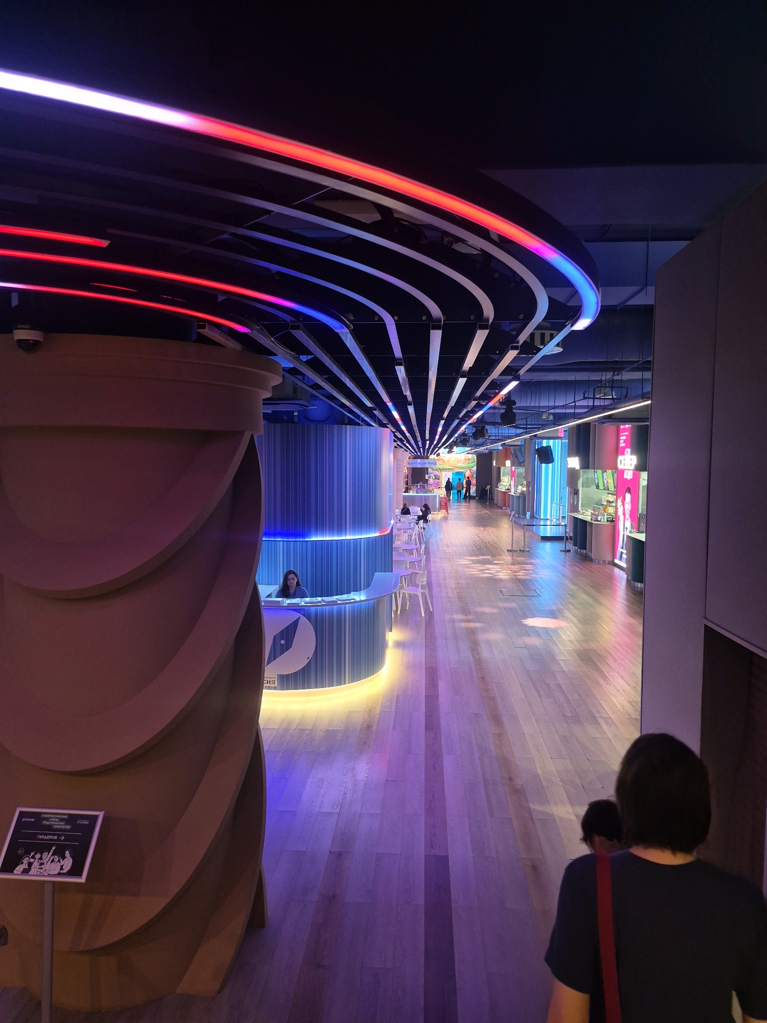 The image captures an interior view of the "Children's Moscow" pavilion at the VDNKh exhibition center in Moscow, Russia. The pavilion is characterized by its futuristic design, featuring sleek, curved architecture illuminated by vibrant LED lighting in shades of red, blue, and purple. The floor is made of polished wood, reflecting the colorful lights.
In the foreground, a large, beige, curved structure dominates the left side of the frame, with a sign that includes an illustration and Russian text. To the right, a reception desk with a woman behind it is visible, with blue and white vertical lighting behind the desk. A circular logo is present on the desk.
The background reveals a wide, illuminated walkway leading into the pavilion. There are several food stalls or exhibition areas visible on the right side of the walkway, with bright signage and displays. People are seen walking along the walkway and in the distance, suggesting a public space with ongoing activity. The overall impression is a modern, dynamic, and visually striking environment designed to engage visitors.