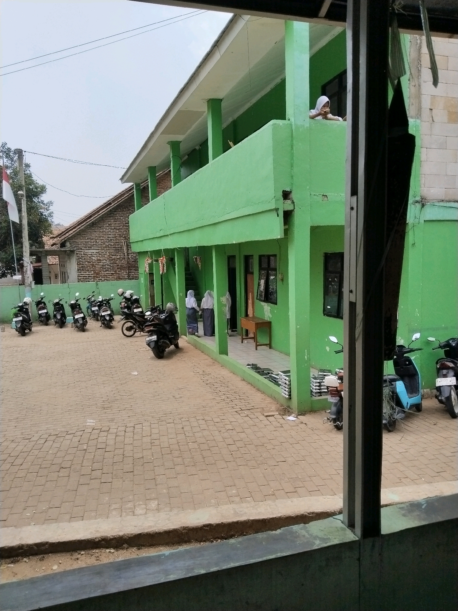 This is a medium-shot, eye-level outdoor photograph taken in the city of Serang, Indonesia, featuring a bright green two-story building, likely a school. In the foreground, a paved courtyard with interlocking bricks is filled with numerous parked motorbikes. To the left, an Indonesian flag on a flagpole waves against a cloudy sky.
The building's ground floor has an open porch area with a simple wooden bench. Several windows are visible on both floors. On the ground floor, stacks of what appear to be learning materials or books are neatly arranged.
On the second floor, a young person wearing a white hijab and a light-colored shirt leans over a green railing, looking downwards. On the ground floor, near the entrance, two individuals wearing similar religious attire stand conversing.
The overall scene depicts a typical day at what appears to be an educational institution in Serang, with students' transportation parked in front of the building. The weather is overcast, suggesting a potentially humid day common in the region. The image is framed by a darker, partially obscured element on the right, suggesting it was taken from within a covered or adjacent structure.