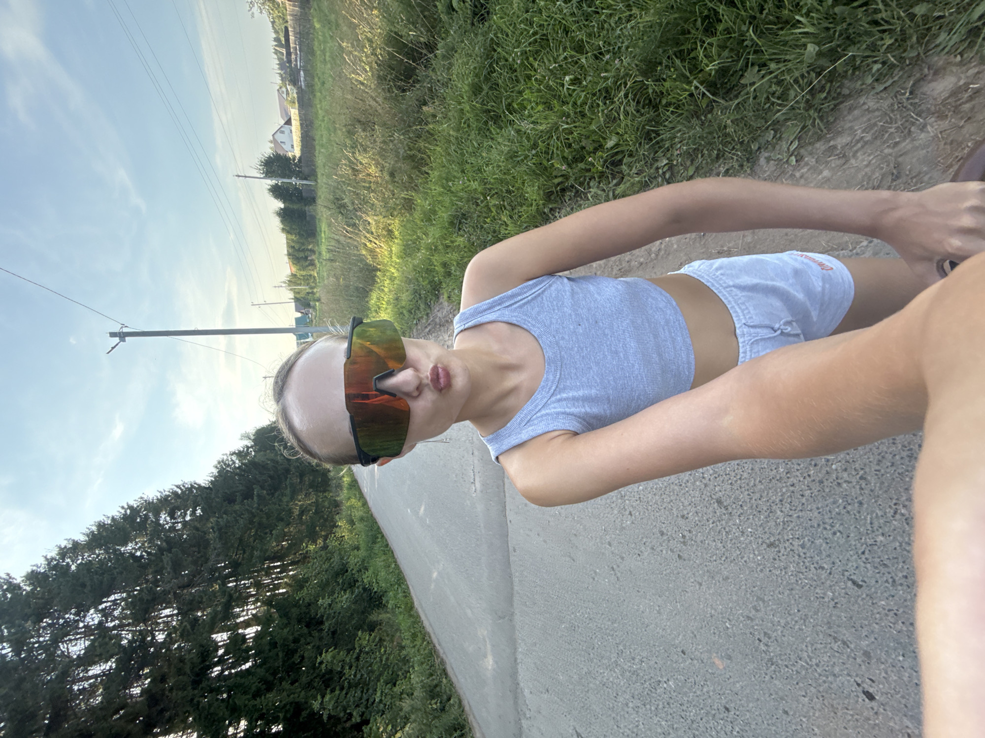A person is standing on a dirt road with a paved path next to it, likely in a rural area given the surrounding greenery and distant buildings. The person is wearing a grey tank top and light-colored shorts, with large, reflective sports sunglasses. They are looking towards the camera and appear to be posing, with their lips pursed. The sun is low in the sky, casting a warm glow and long shadows, indicating it is either early morning or late afternoon. The weather appears to be clear and pleasant. The scene is set with a backdrop of lush green grass, trees, and a blue sky with some wispy clouds. There are power lines and poles visible, suggesting the presence of infrastructure in the area. The location context provided is Подшивалово, Russia, which aligns with the rural scenery. There is no visible text in the image.