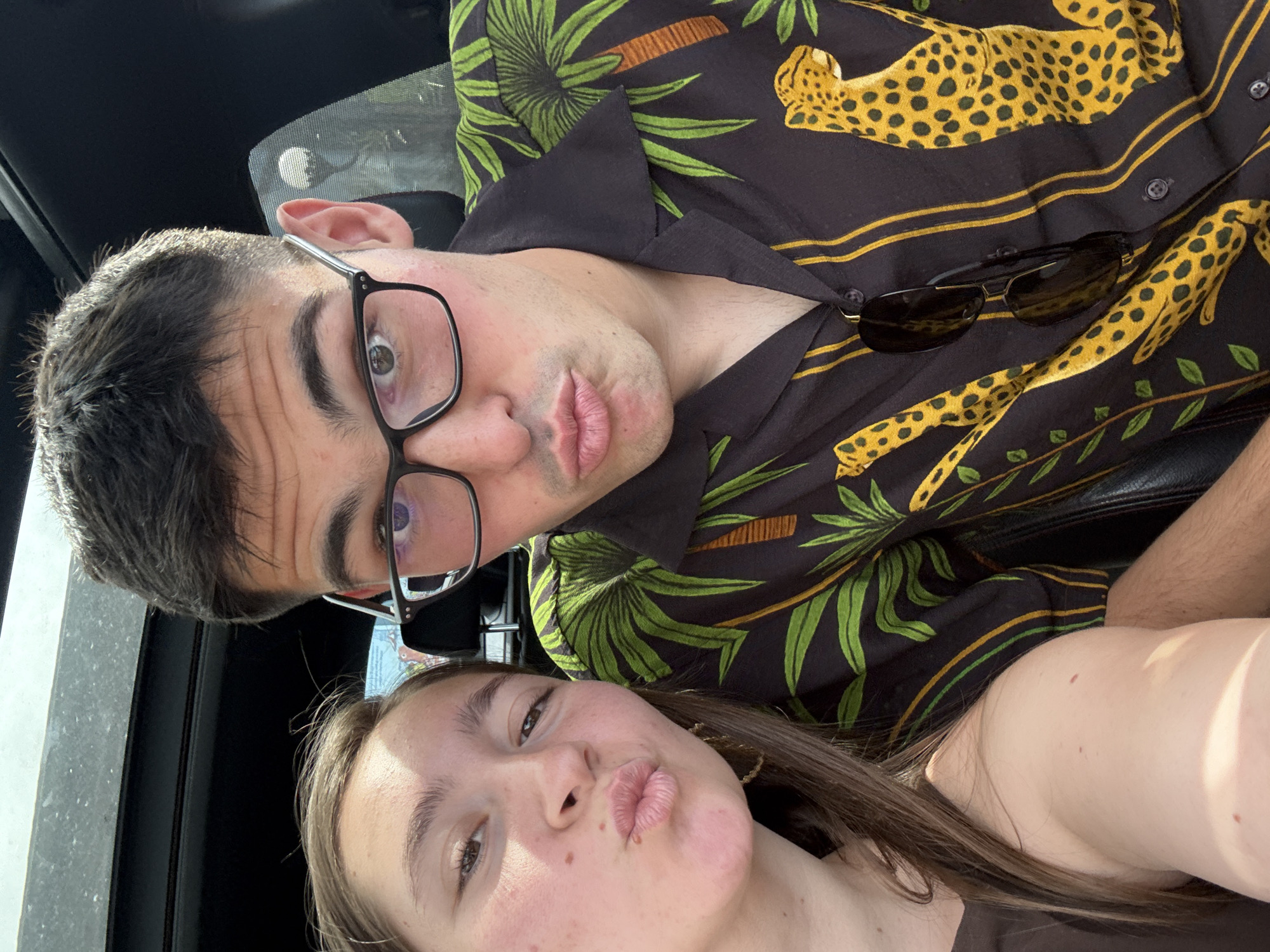 The image, taken in Radinovo, Bulgaria, captures a close-up selfie of two individuals, a male and a female, within what appears to be the interior of a vehicle. The perspective is from a low angle, looking upwards at the subjects, who are positioned close together in a playful pose. Both have their lips slightly puckered.
The male subject, prominent in the upper and central part of the frame, wears black-framed glasses and has short, dark hair. He is dressed in a black shirt adorned with a distinctive pattern of green palm leaves and yellow cheetah or leopard figures. A pair of dark sunglasses is casually tucked into the collar of his shirt.
The female subject is in the lower section of the frame, featuring long brown hair and wearing a dark, possibly brown, top. Her expression mirrors the male's, contributing to the casual and lighthearted mood of the photograph.
The scene is brightly illuminated by natural daylight, suggesting the picture was taken during daytime, likely under sunny conditions, as indicated by the clear reflections in the male's glasses and the general brightness. No discernible text is visible on clothing, objects, or in the background. The overall impression is one of a shared, affectionate moment.