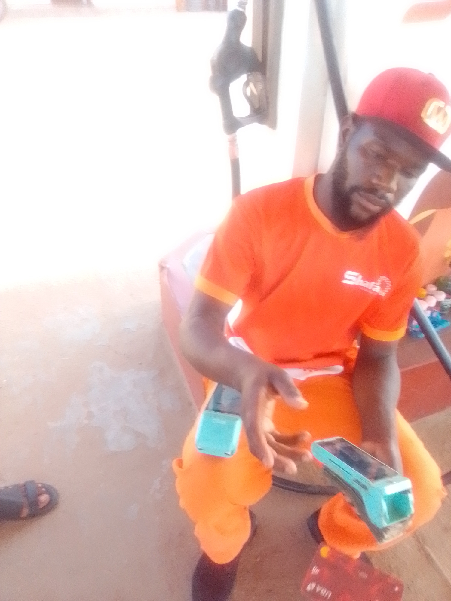 A man wearing an orange shirt and orange pants, with a red cap, is seated in what appears to be a gas station in Kwadon, Nigeria. He is holding two electronic payment devices, one in each hand. The man has a beard and appears to be looking down at the devices. The background shows a gas pump nozzle and a concrete-like surface. There is a red card on the ground near his feet. The lighting suggests it is daytime.