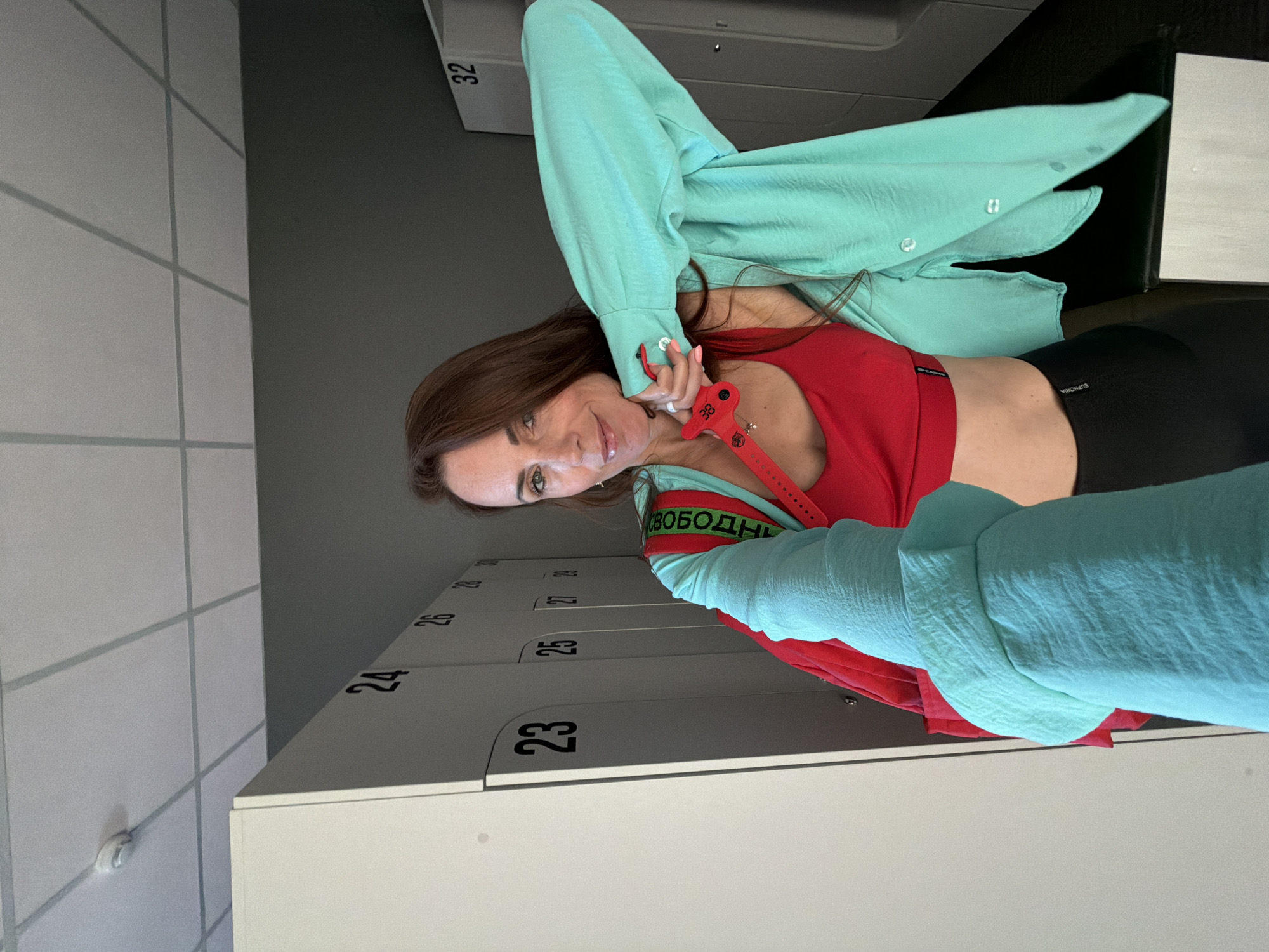 A woman with long brown hair is standing in a locker room, posing for a selfie. She is wearing a red sports bra, black leggings, and an unbuttoned turquoise shirt. A red bag with a green strap is slung over her shoulder. The locker room has white lockers with numbers on them, such as 23, 24, 25, 26, 27, 28, 29, 32. The ceiling is made of acoustic tiles. The overall environment is clean and well-lit. There is no indication of the city of Birobidzhan, Russia in the image.