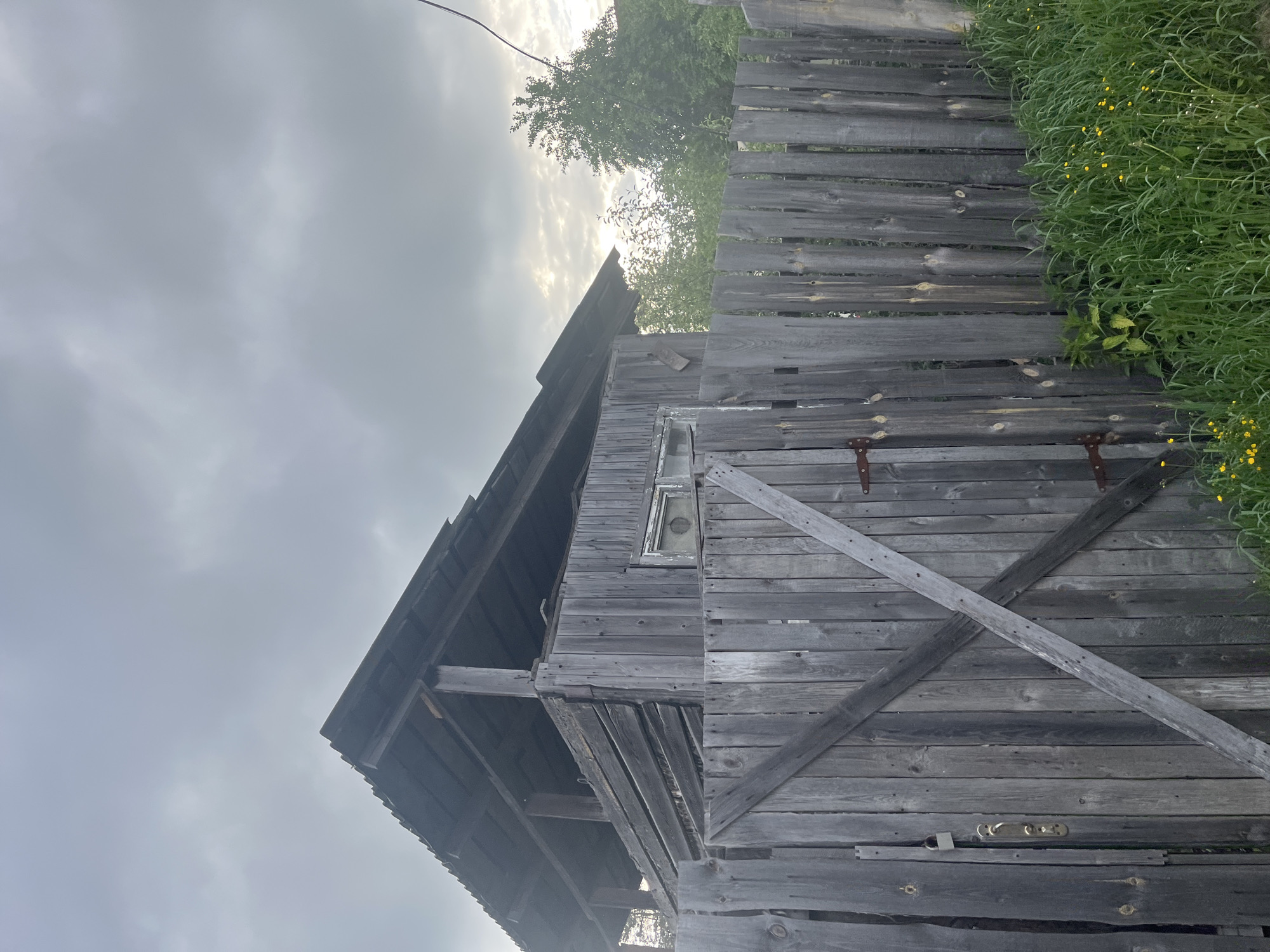 The image shows an old, weathered wooden structure, likely an outbuilding or shed, in Chaikovsky, Russia. The wood is gray and aged, with visible weathering and some deterioration. The structure has a sloped roof and a small window. A wooden fence stands to the right, with greenery and some small yellow flowers growing nearby. The sky is overcast. The scene appears to be outdoors during the day.