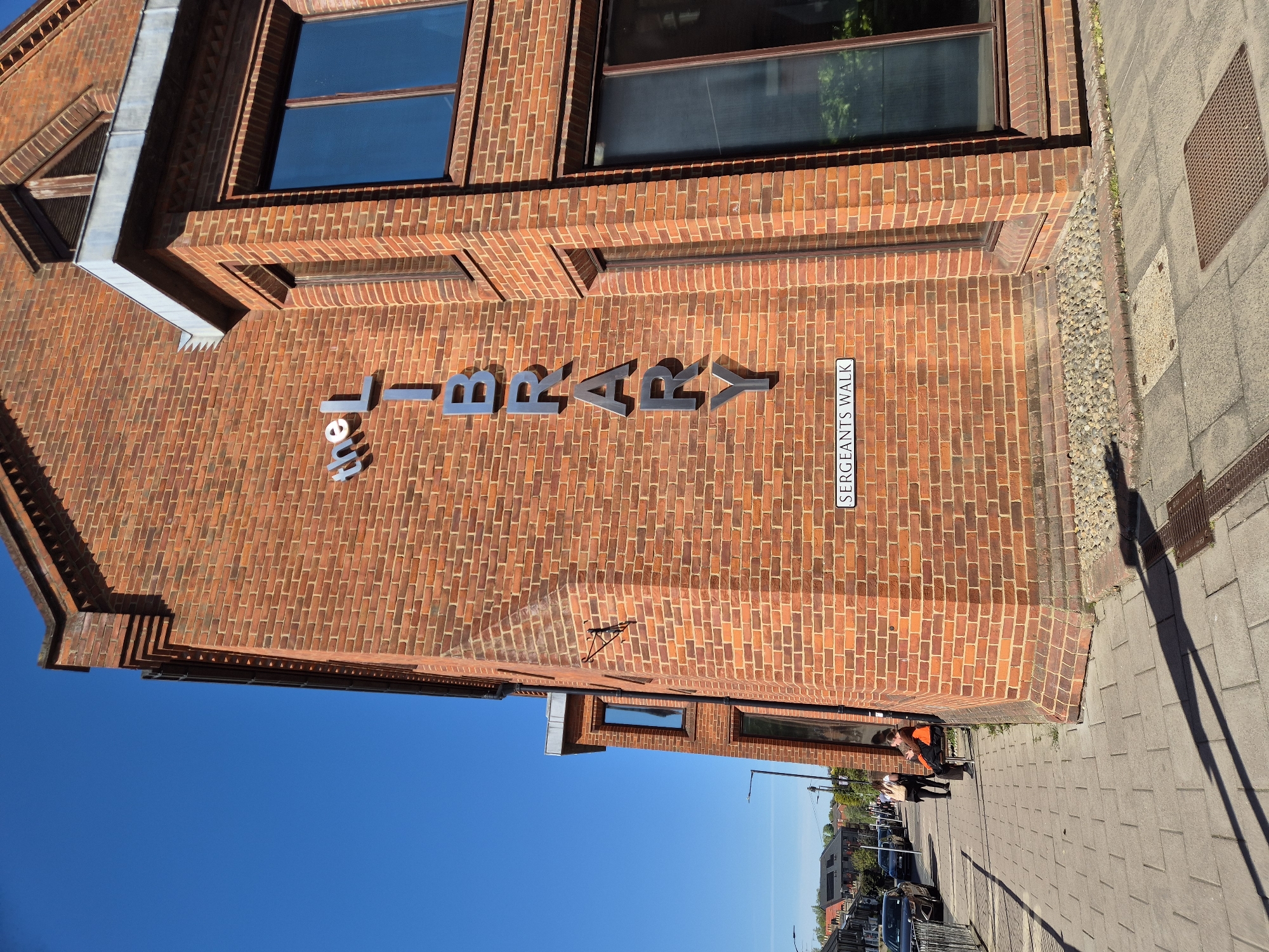 A brick building is partially shown in an upward angle, with its signage indicating it is "The Library" on Sergeants Walk. The architecture features brickwork with window openings. A paved walkway runs alongside the building's exterior, suggesting an urban environment. Distant trees, light poles, and a row of parked cars are visible in the background, indicating a street scene, suggesting the location is in West Suffolk, United Kingdom.