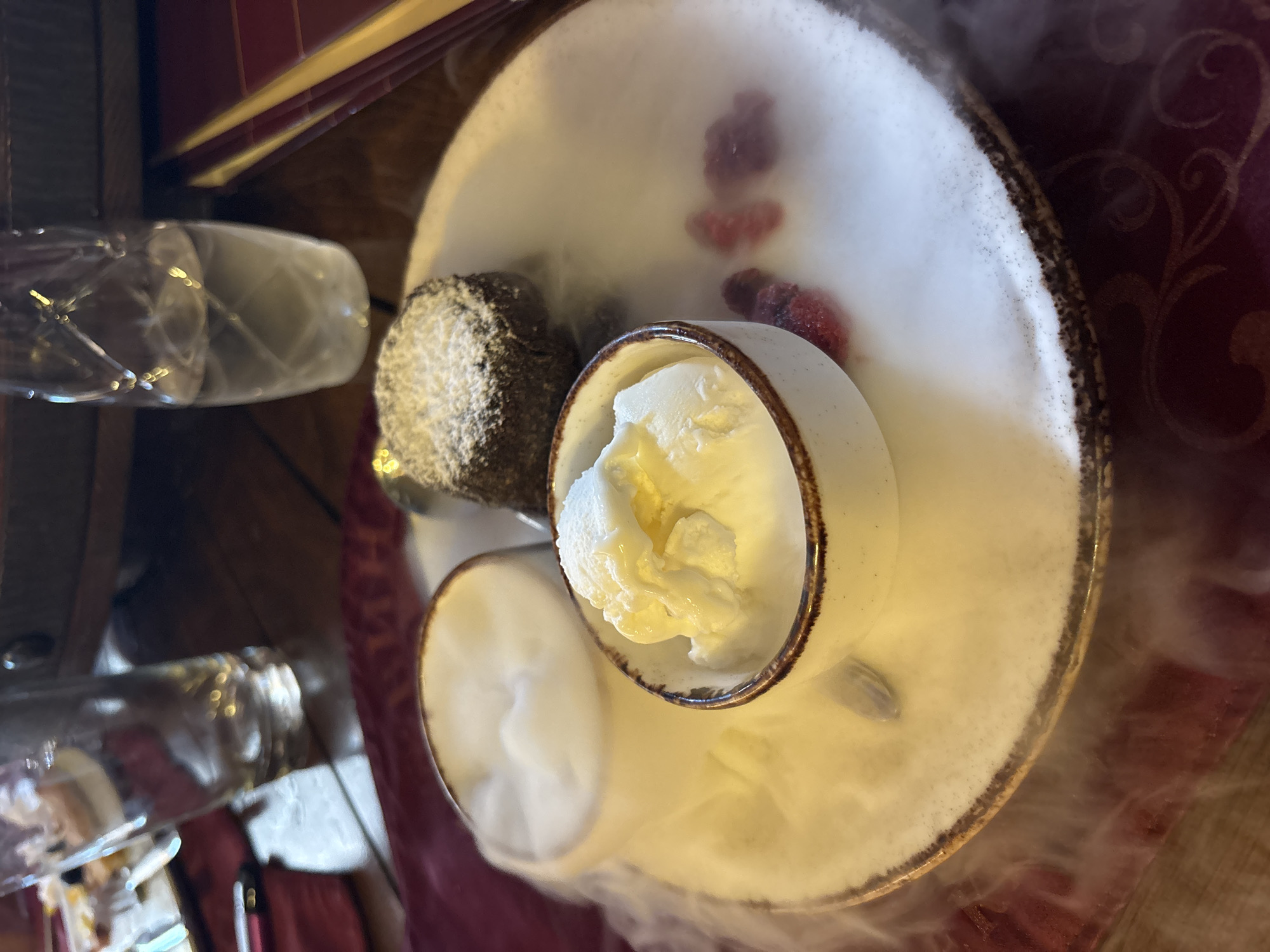 A table is set for a dessert, featuring a chocolate lava cake dusted with powdered sugar, served with a scoop of vanilla ice cream and a side of what appears to be whipped cream or mousse, all presented on a white plate. Wisps of dry ice smoke surround the desserts, creating a dramatic presentation. The setting appears to be a restaurant, with a wooden table and glassware visible in the background. The cuisine is European, likely Bulgarian, given the location in Lukovit, Bulgaria. The image captures a moment of indulgence, with a focus on the rich textures and presentation of the dessert.