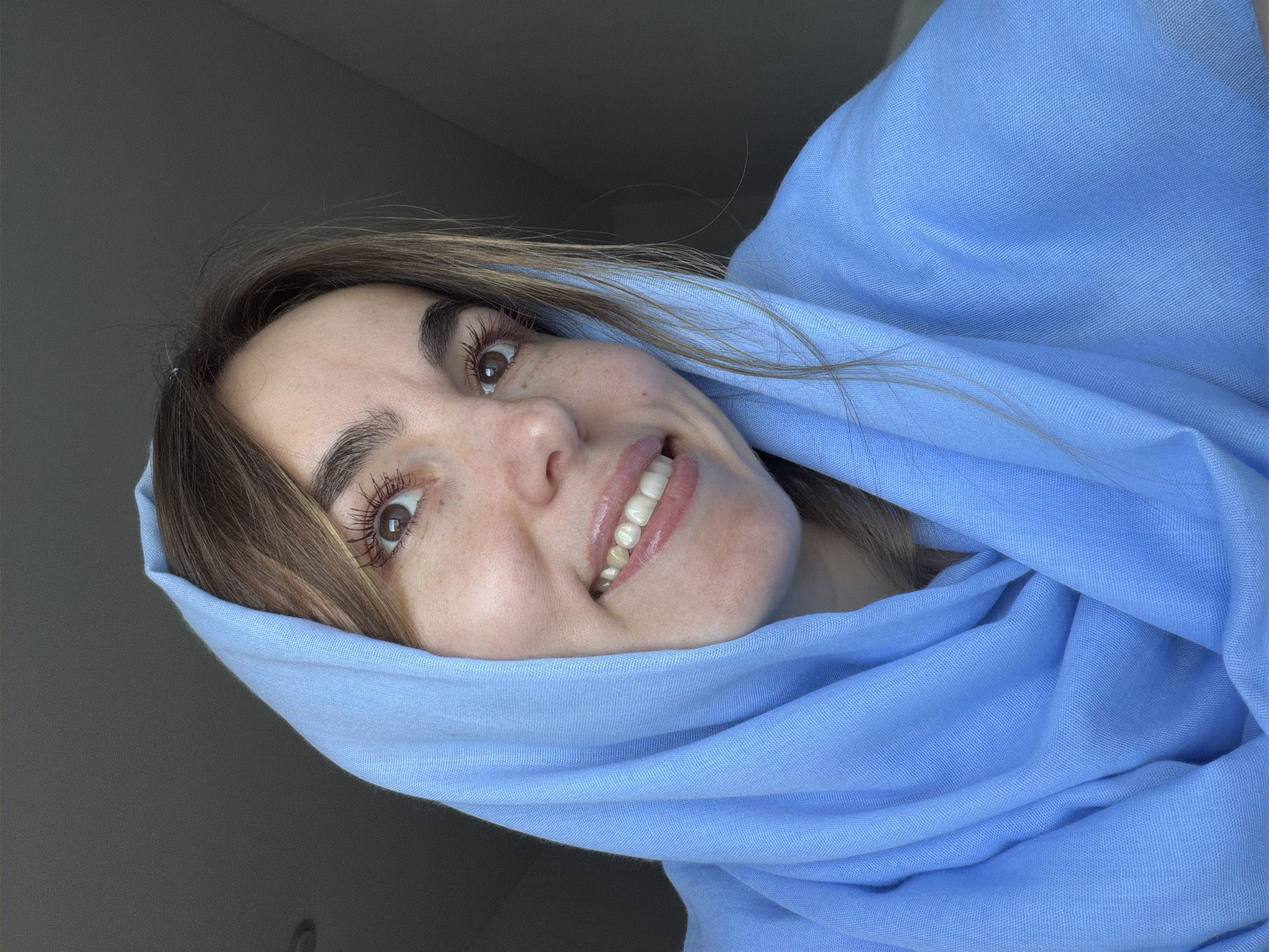 A woman with light brown hair and fair skin smiles broadly at the camera, wearing a blue headscarf. The background appears to be a gray wall. It is not possible to determine if the image was taken in Sarapul, Russia, based on the visual content.