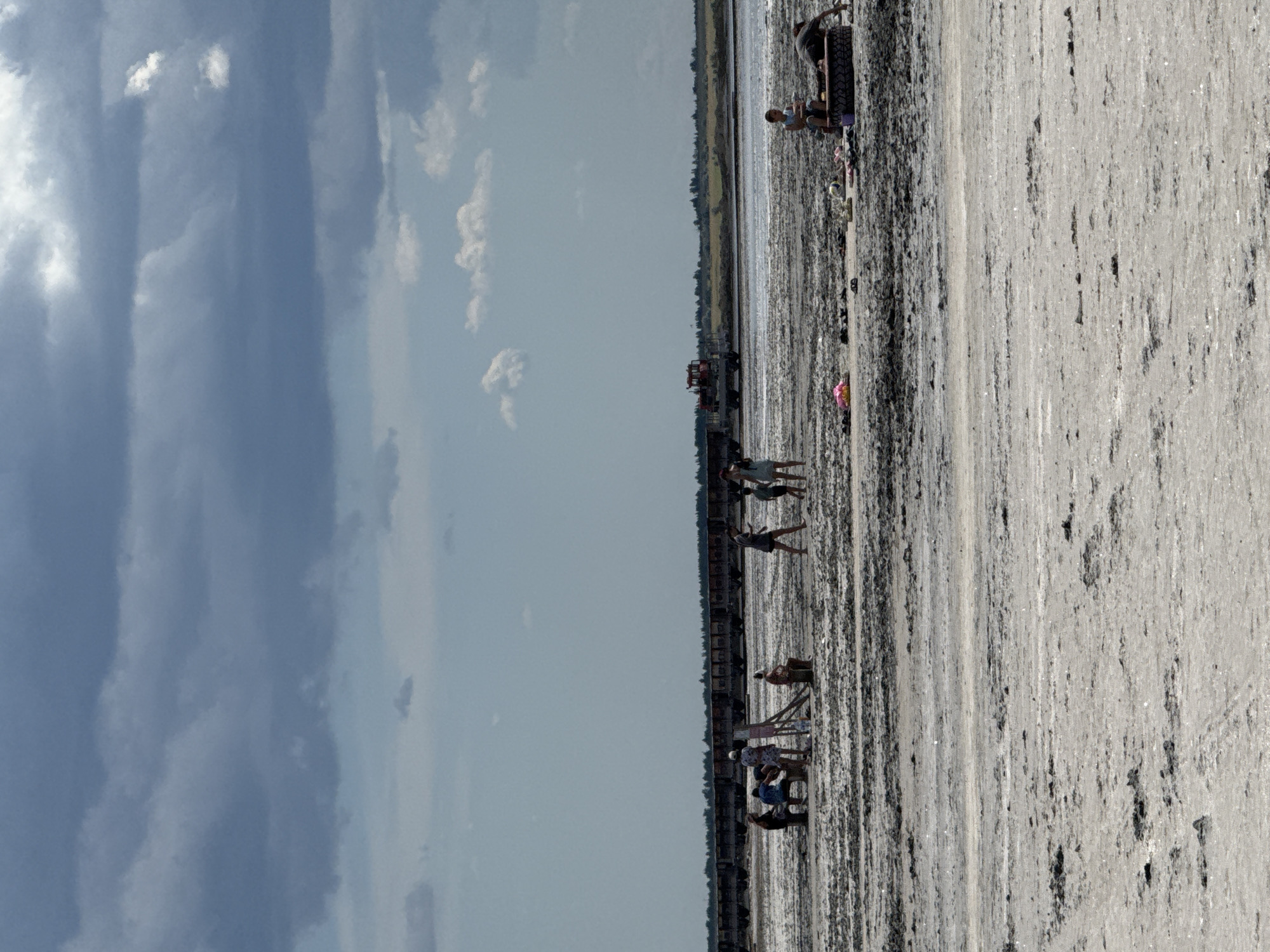 The image depicts a wide, flat expanse of salt flats, likely located in Bursol, Russia, under a dramatic, overcast sky filled with dark, heavy clouds. In the distance, a long train of rail cars stretches across the horizon, with a red locomotive visible on the right side. Several scattered groups of people are present on the salt flats. In the left middle ground, a group of four individuals are gathered, with one person appearing to paint or draw on an easel. Further to the right, a group of three people are visible, and in the far right middle ground, two individuals are relaxing on what appears to be a large tire. Scattered on the salt flats are various personal items including shoes and bags. The ground itself is a textured mix of light-colored salt and darker, muddy patches, suggesting a recently dried-up body of water.