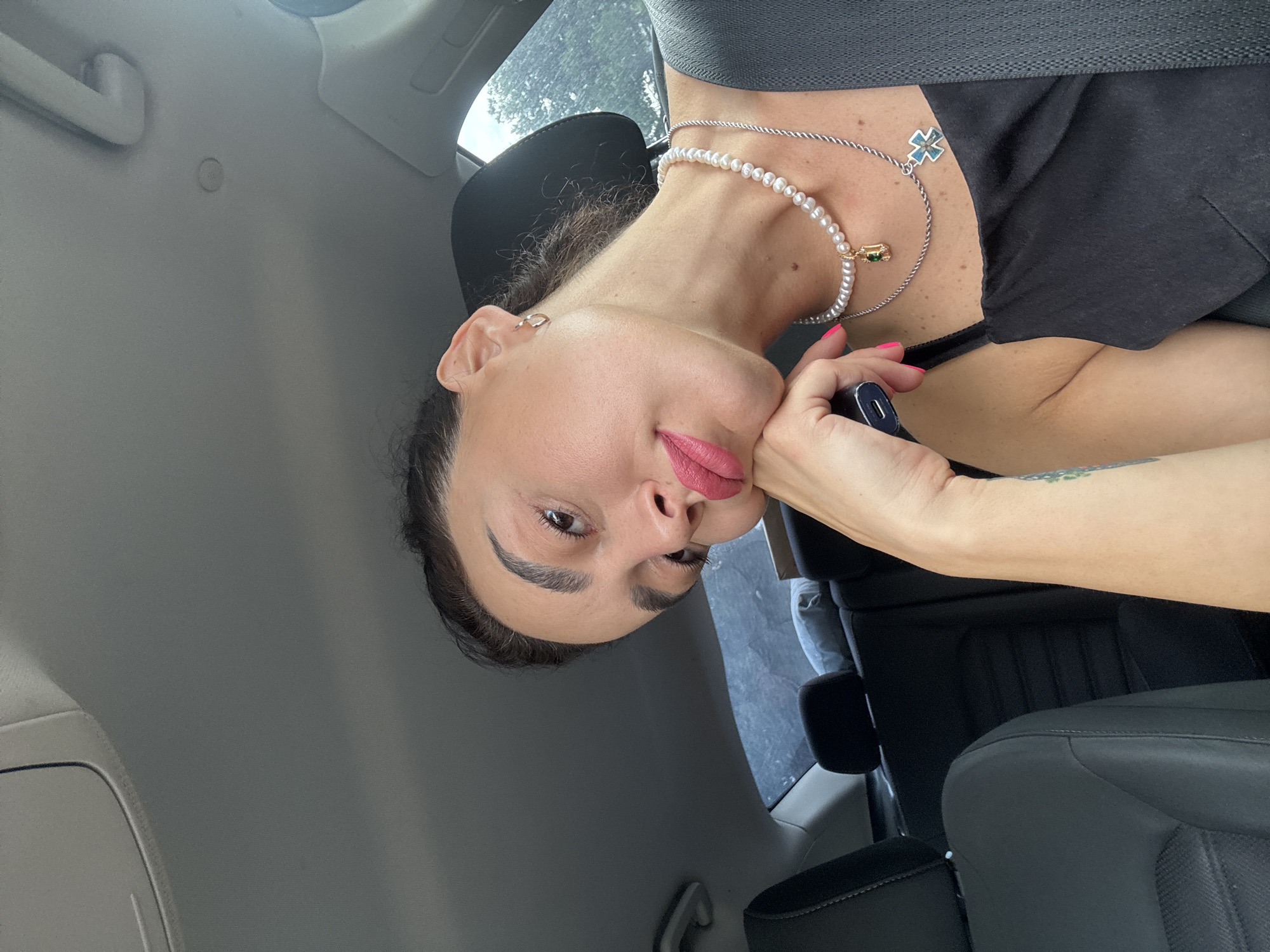 The image shows a woman sitting in the passenger seat of a car. She is leaning her head on her hand, with her eyes looking down. She is wearing a black top, a pearl necklace, and earrings. Her fingernails are painted pink. In the background, the interior of the car is visible, including the seats and the ceiling. The woman is wearing makeup, including lipstick. The scene appears to be in an urban environment. There is no specific indication of the location being Tbilisi, Georgia.