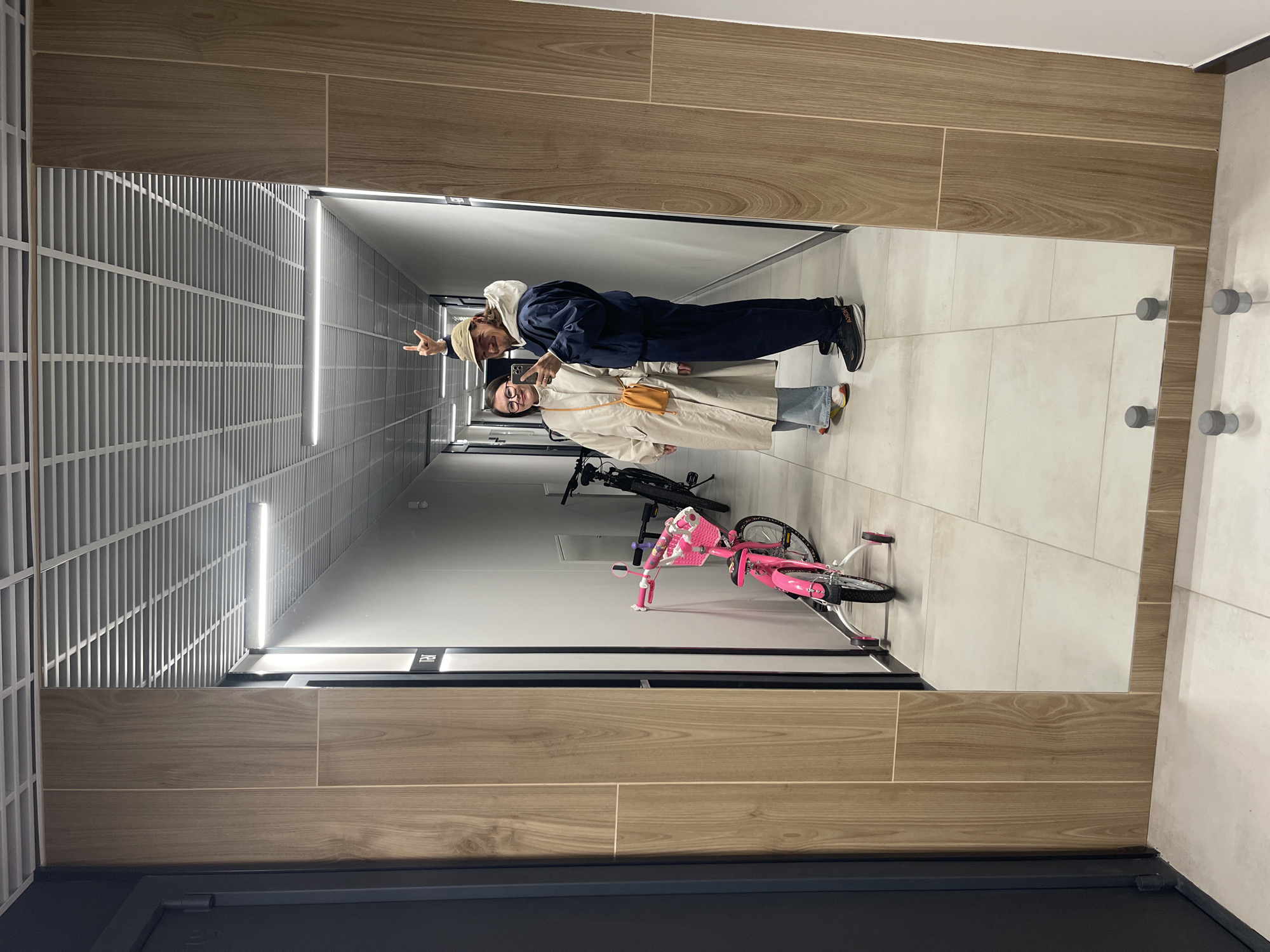 The image captures a reflection of two individuals in a corridor. The corridor is well-lit with recessed fluorescent lights, and the walls are a mix of white and textured panels. A pink bicycle is parked against the wall further down the corridor.
One individual, on the left, is wearing glasses and a beige coat. The other individual, on the right, has a baseball cap on and is wearing a navy blue jacket and pants. They appear to be posing for a photo or taking a selfie. They are both smiling at the camera in the reflection.
The corridor appears to be in a residential or commercial building, as indicated by the presence of bicycles and the design of the hallway. The location in the city of Unknown, Unknown is not discernible from the visual data in the image.
